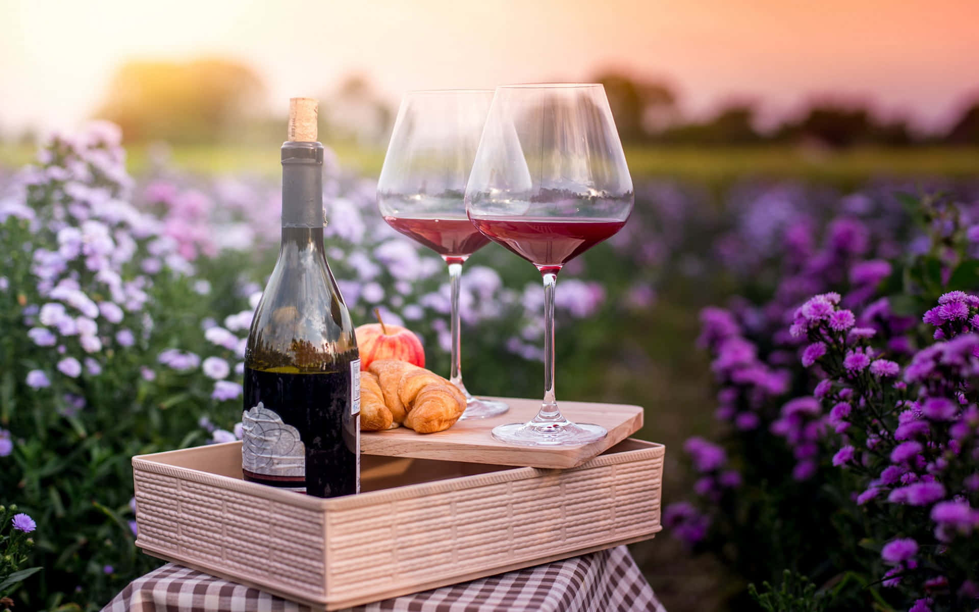 Wine 桌布 壁紙 — 在紫色花田日落背景下，編織托盤上擺放著酒杯、可頌和一瓶紅酒的野餐佈置。