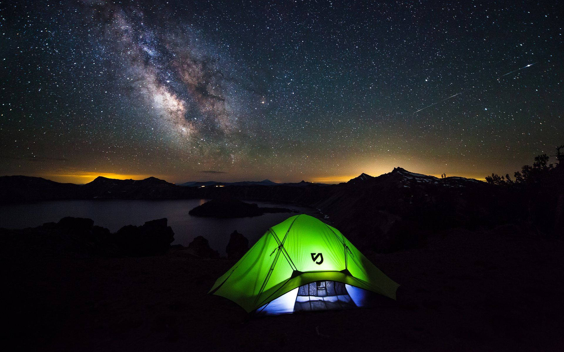  壁紙 — 在繁星點點的夜空下，一個發光的綠色帳篷，銀河在黑暗的湖泊和山脈上空拱起。