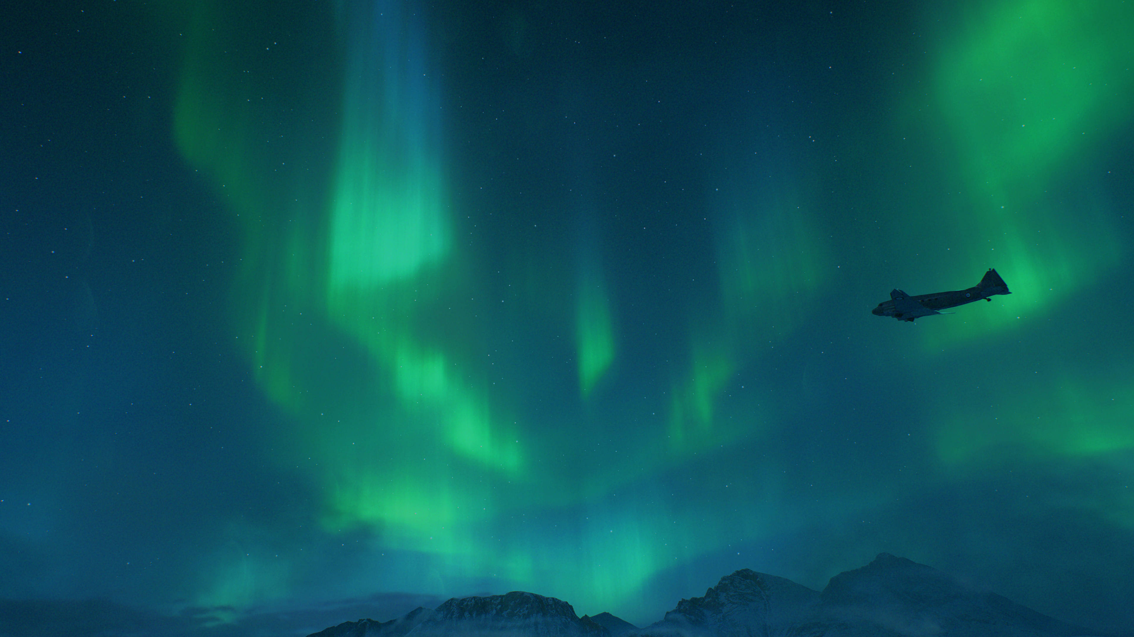 aurora borealis 桌布 壁紙 — 在繁星點點的夜空下，一架飛機的剪影飛過在雪山上方舞動的鮮綠色北極光。