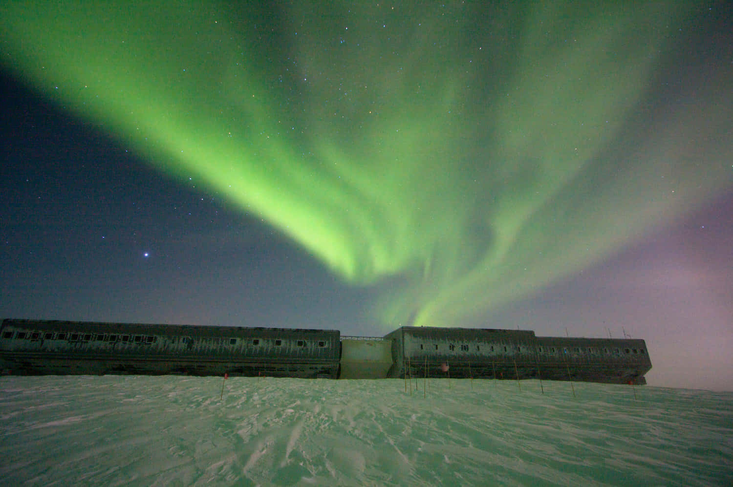 Antarctica 桌布 壁紙 — 在繁星點點的夜空下，一道充滿活力的綠色極光在積雪覆蓋的廢棄火車車廂上方舞動。