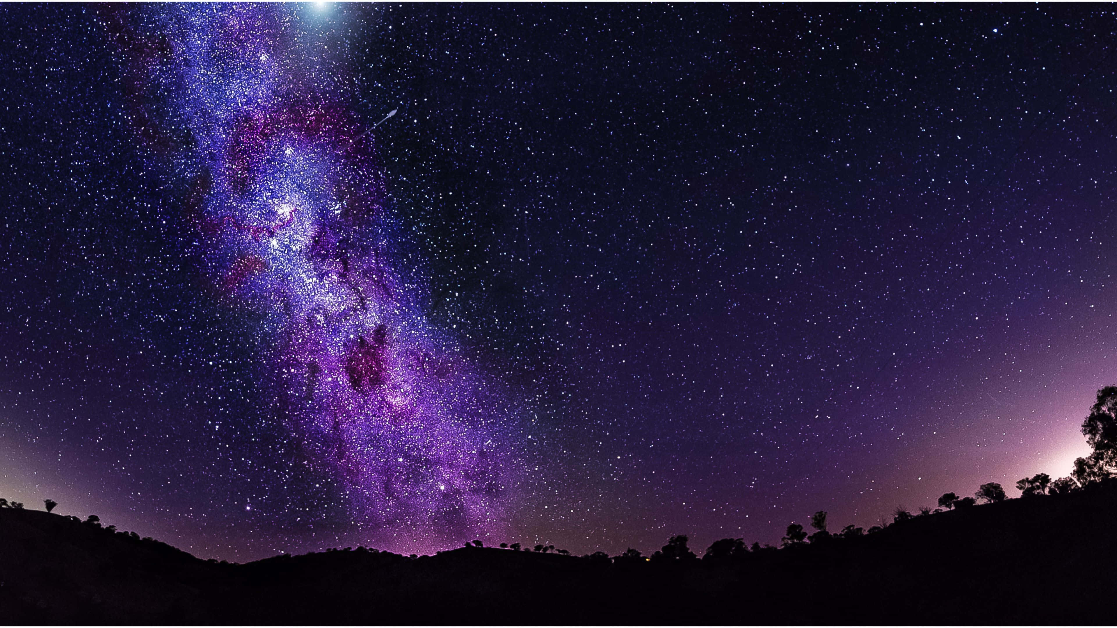 銀河 桌布 壁紙 — 在繁星點點的夜空下，一道壯麗的銀河在黑暗的山丘剪影上方閃耀著鮮豔的紫色和藍色色調。