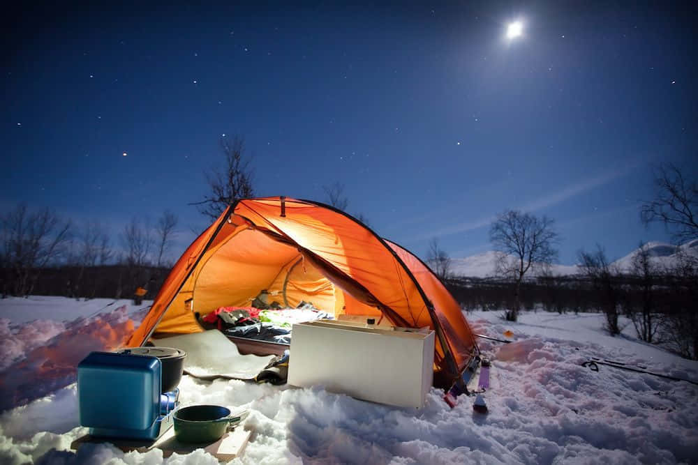Winter Camping 桌布 壁紙 — 在繁星點點的夜空下，一頂發光的橙色帳篷坐落在雪地荒野中，頭頂上有一輪明亮的月亮。