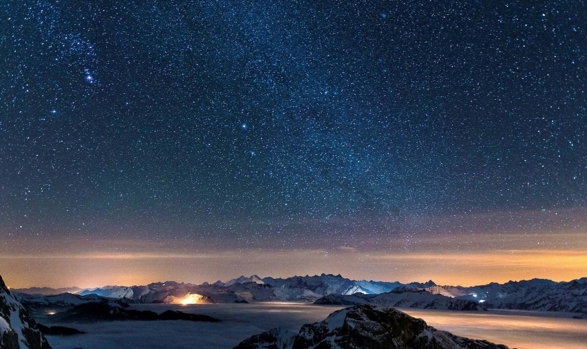 Starry Sky 桌布 壁紙 — 在繁星點點的夜空下，積雪的山峰伴隨著發光的極光和遠處的燈光。