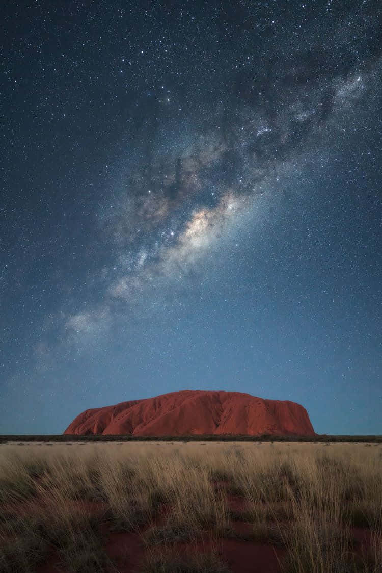 Rock 桌布 壁紙 — 在繁星點點的沙漠夜空下，銀河壯麗地拱起在標誌性的紅色巨石烏魯魯之上。