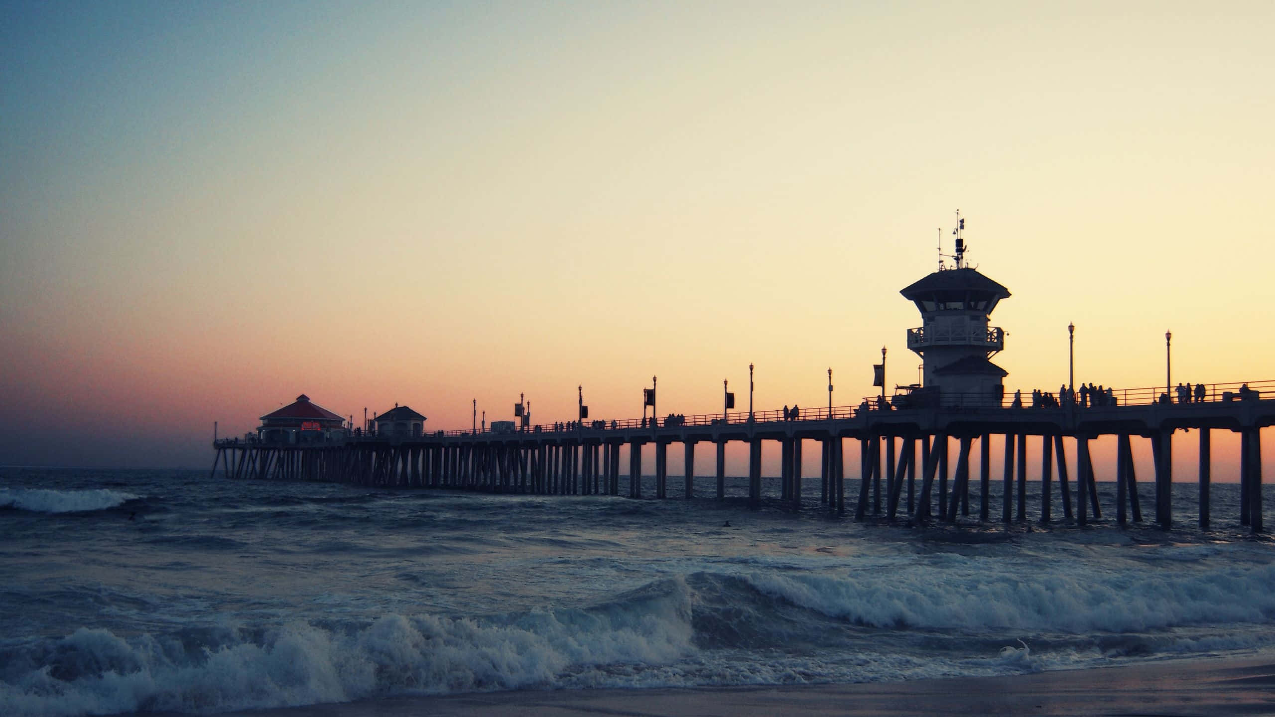 Pier 桌布 壁紙 — 在翻滾的海浪之上，以 vibrant 的日落天空為背景，呈現出燈塔和人們剪影的碼頭。
