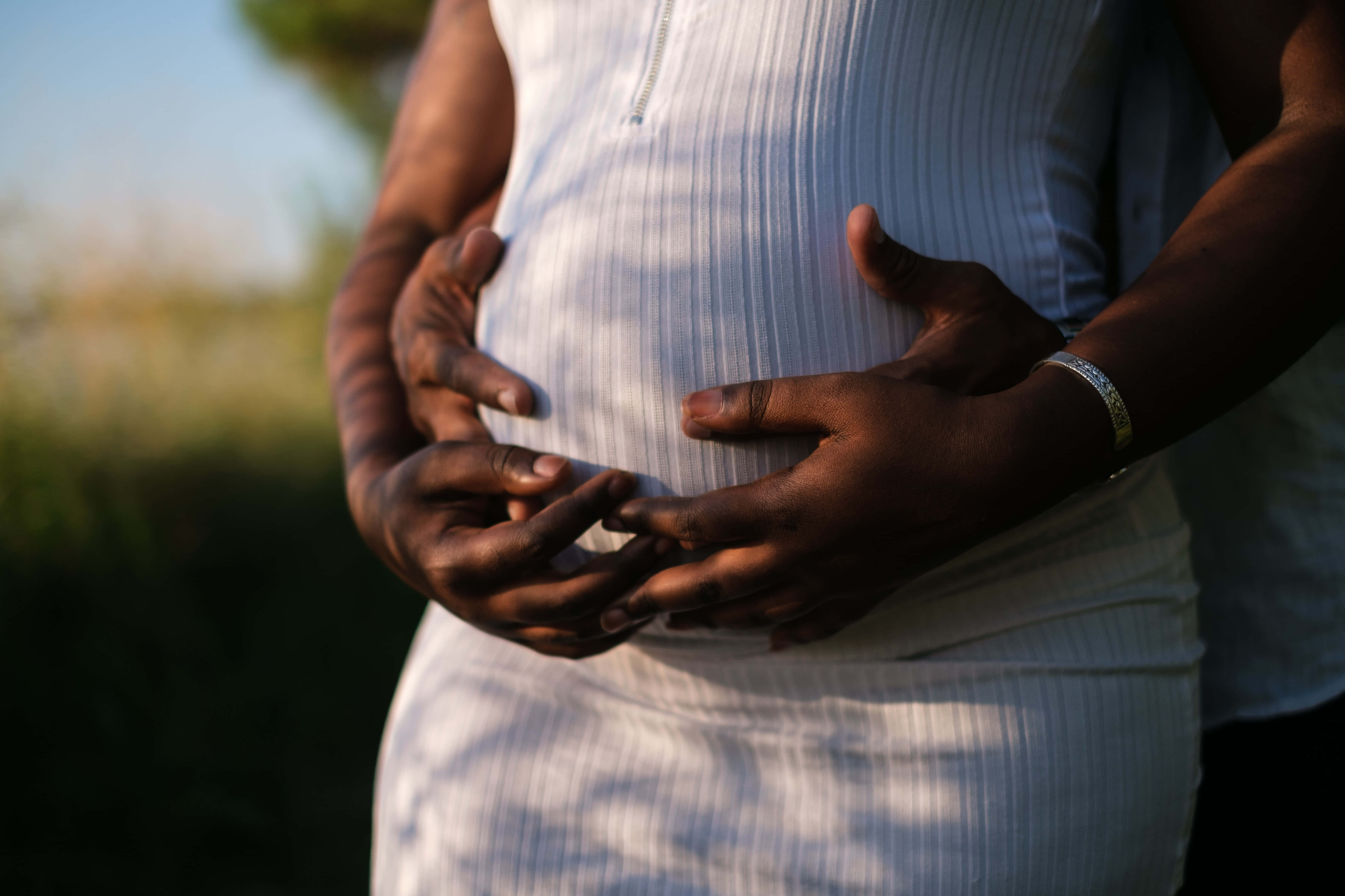 Pregnant 桌布 壁紙 — 在自然戶外環境中，斑駁的陽光下，兩隻手輕柔地捧著懷孕的肚子，一個溫柔的時刻。