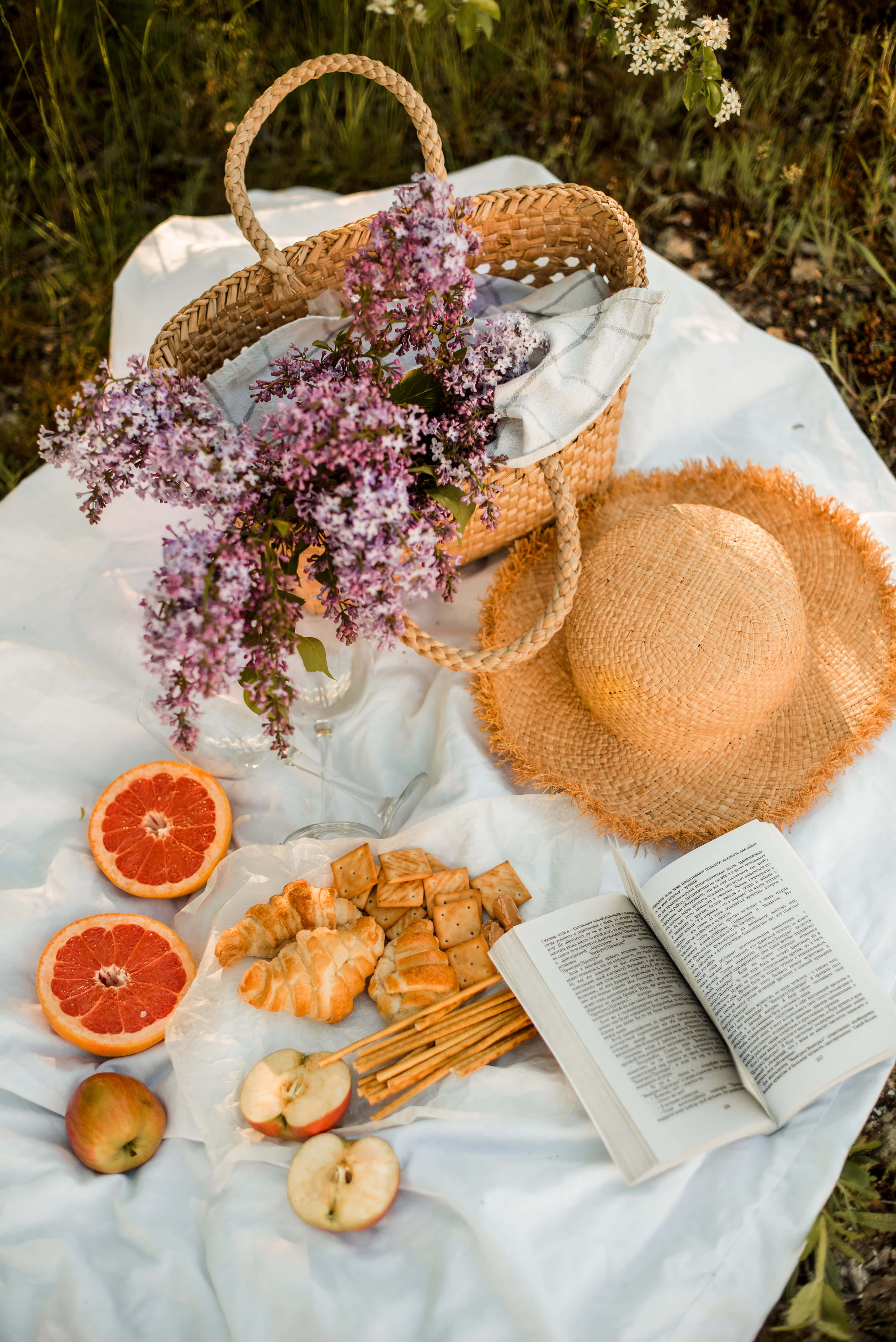 picnic 桌布 壁紙 — 在草地上白色毯子上擺放著丁香花、草帽和切片水果的寧靜野餐佈置。