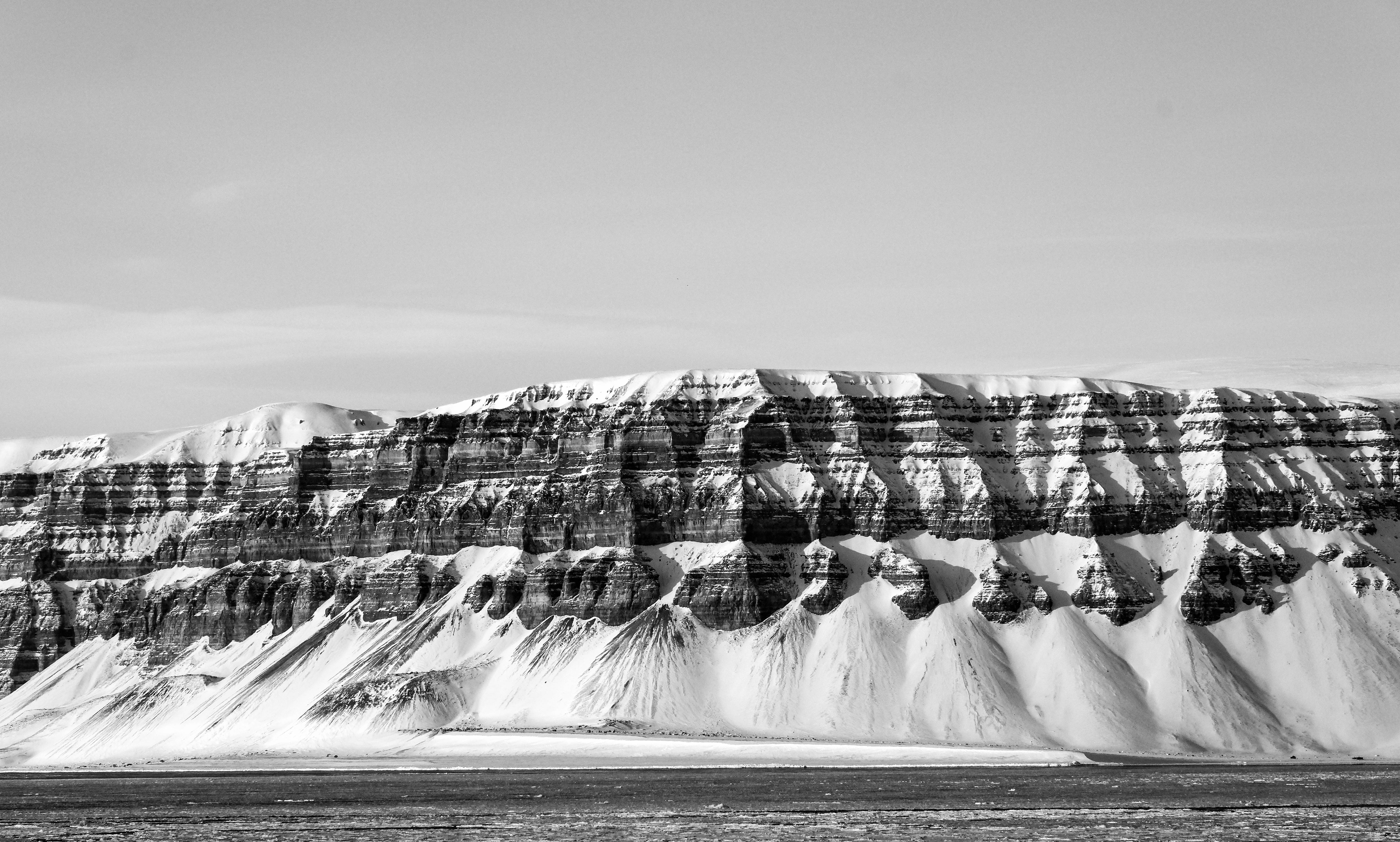 Svalbard 桌布 壁紙 — 在蒼白天空下，雪塵覆蓋的層狀懸崖的鮮明黑白景觀。