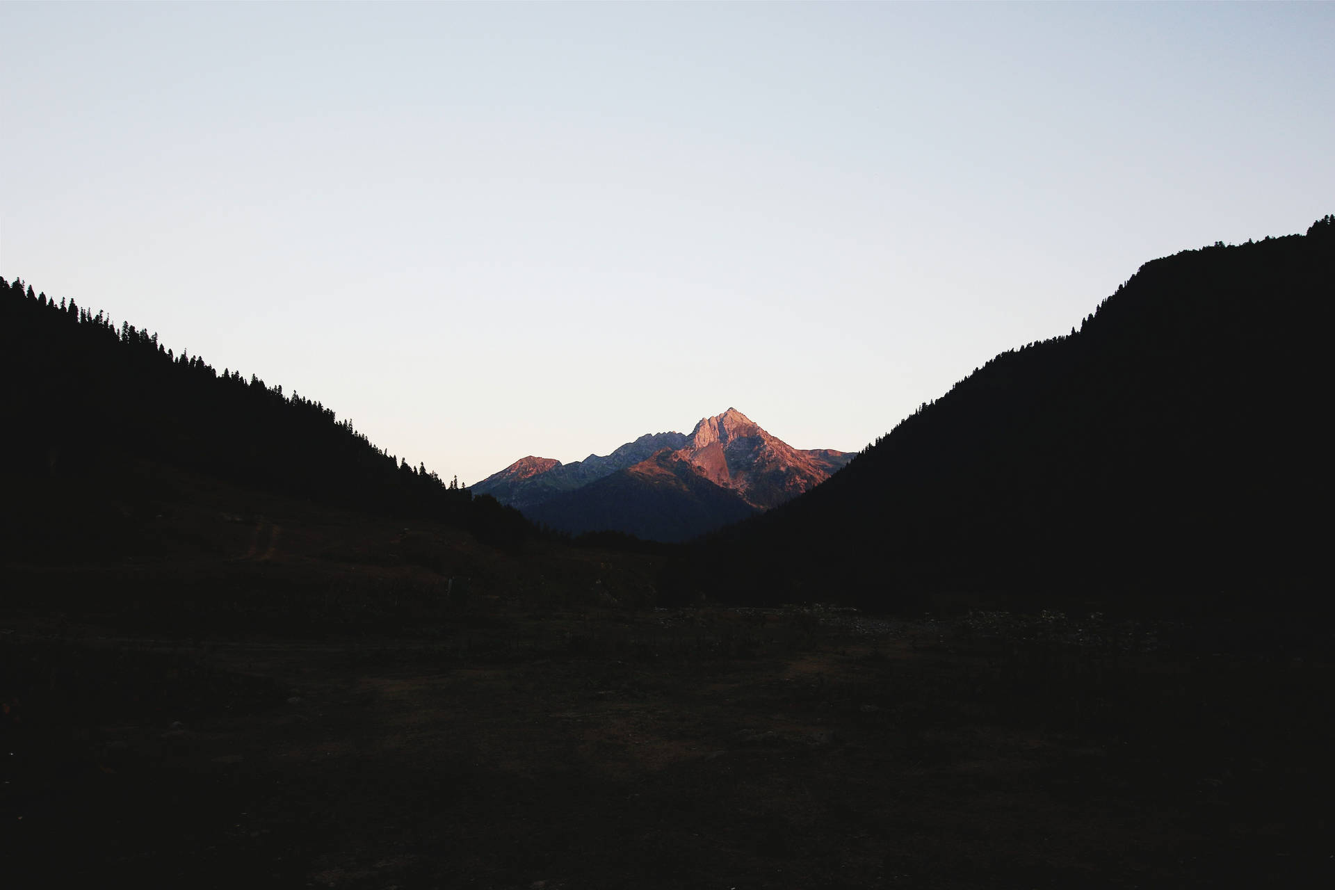 一座山峰沐浴在日落光芒中，被黑暗的森林山丘環繞。 — 山脈 壁紙