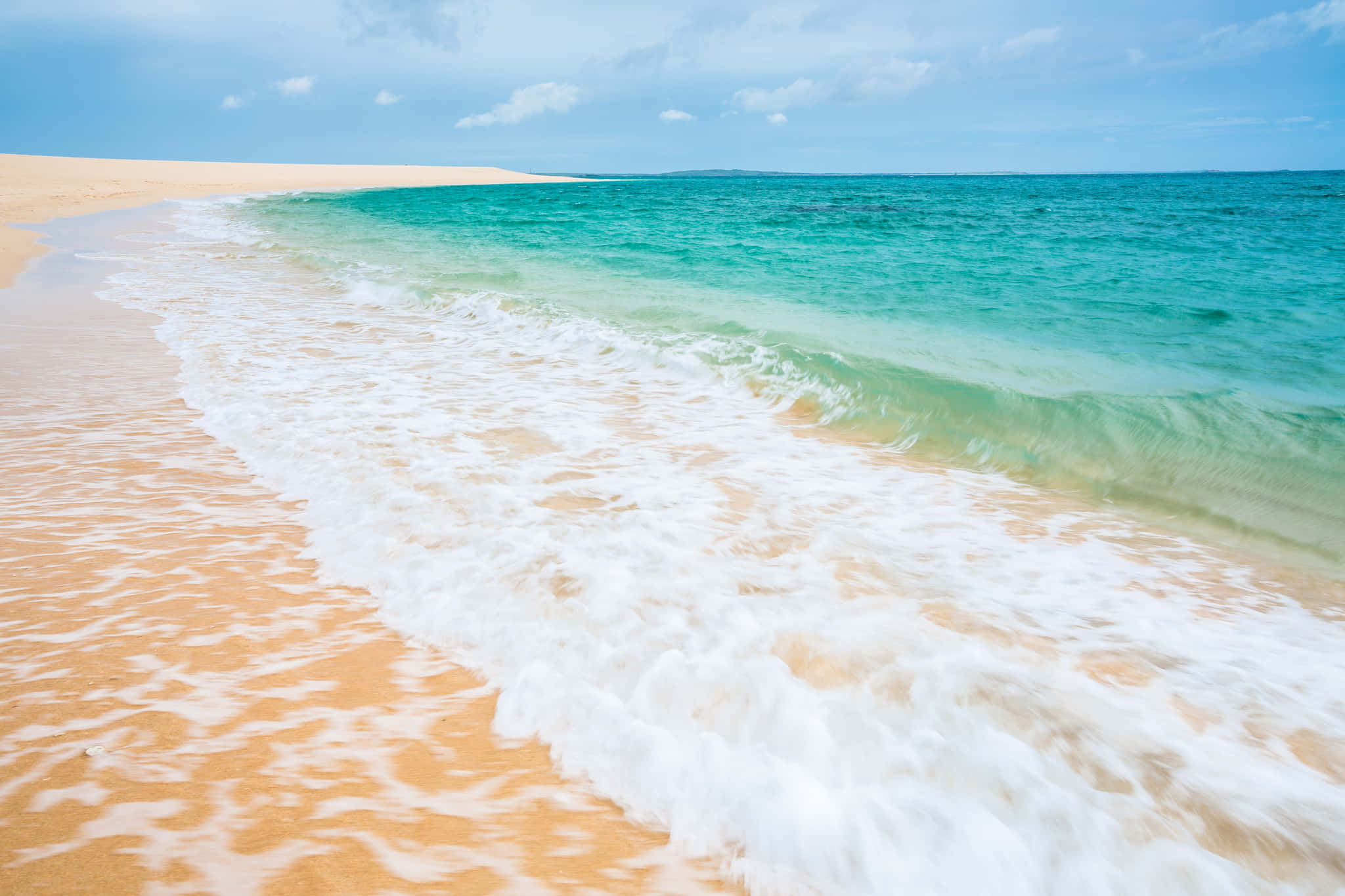 Beach Sand 桌布 壁紙 — 在蔚藍天空和零星雲朵下，碧綠色的海浪拍打著金色沙灘。