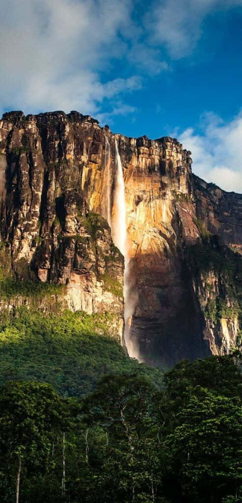 Venezuela 桌布 壁紙 — 在蔚藍天空與零星白雲下，一座雄偉的瀑布從高聳的懸崖峭壁傾瀉而下，四周環繞著茂密的綠色叢林。