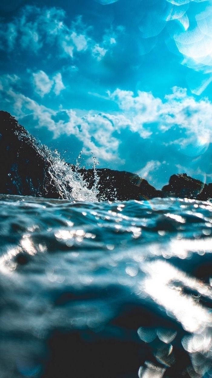 桌布 壁紙 — 在藍天白雲下，海浪拍打黑色岩石的動態水下視角。