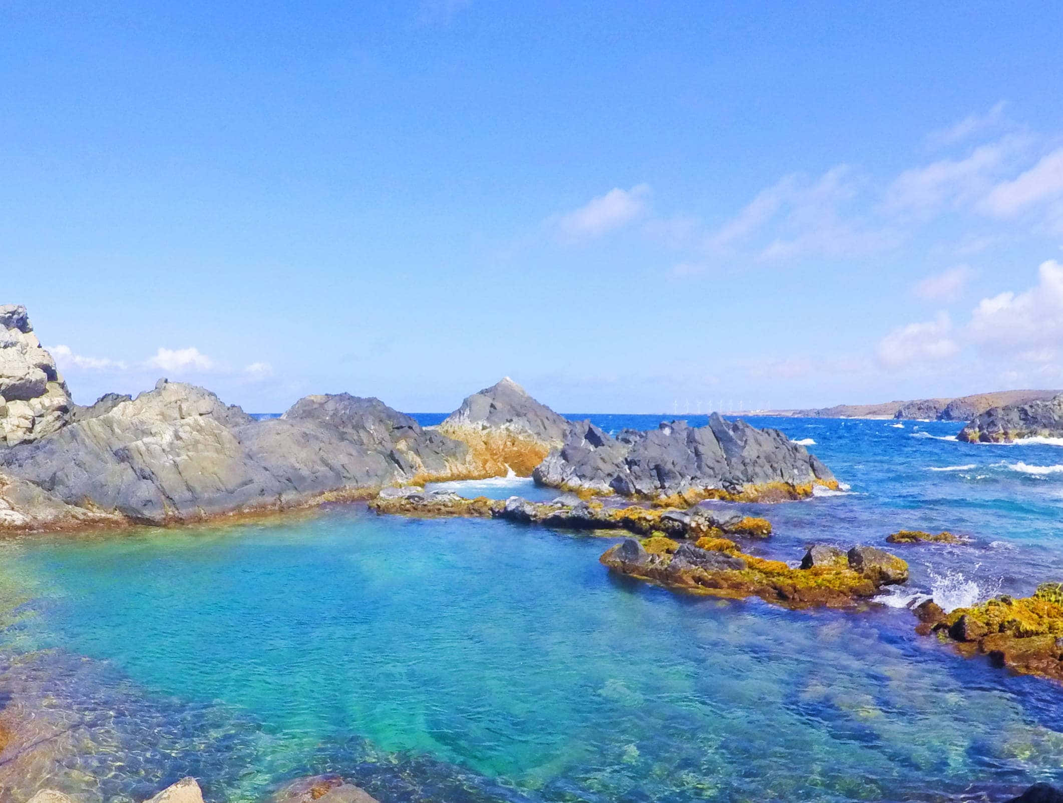 Aruba 桌布 壁紙 — 在藍天白雲下，清澈的碧綠海水拍打著粗糙的灰色岩石。
