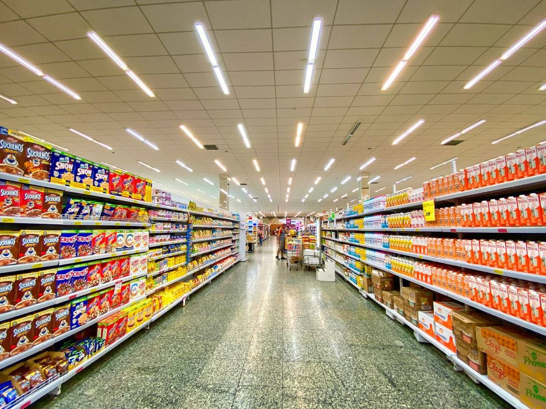 Shopping 桌布 壁紙 — 在螢光燈下，一條光線充足的超市通道，色彩繽紛的零食貨架延伸至遠方。