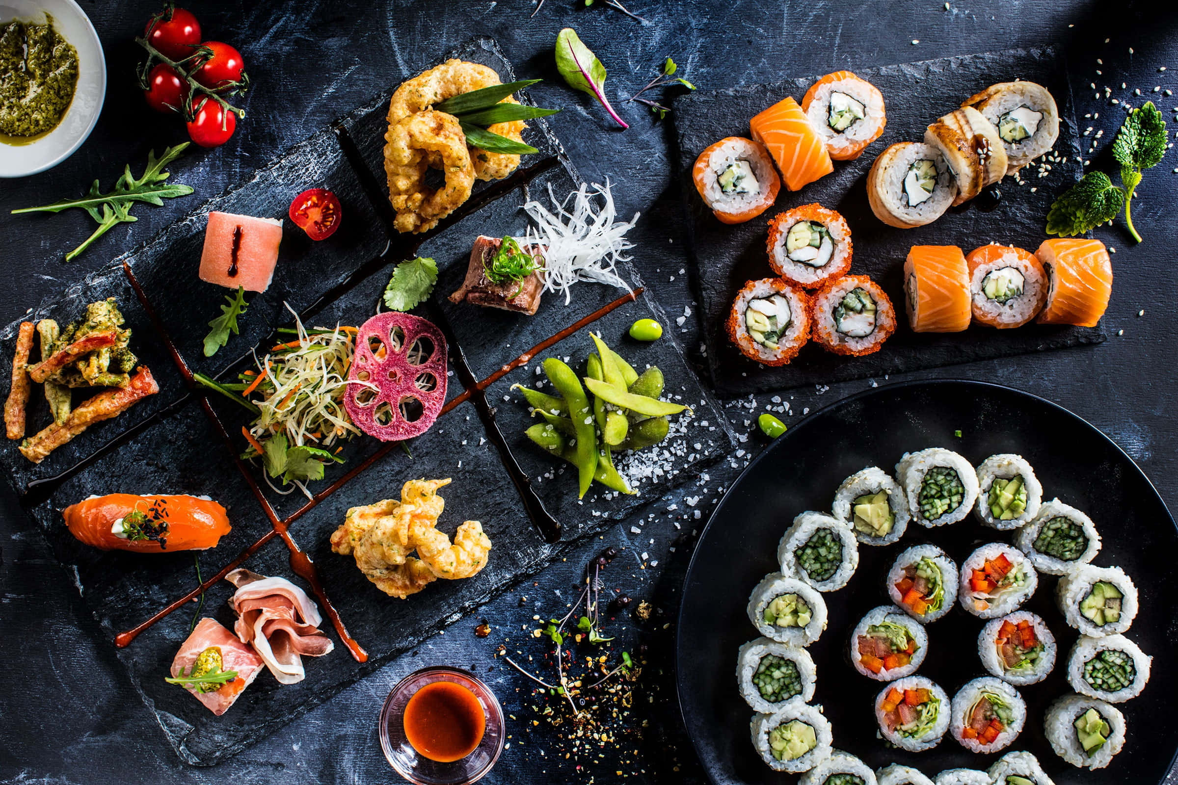 Food Photography 桌布 壁紙 — 在質感黑色背景上，深色石板盤上精心擺放著豐盛的壽司卷、刺身和天婦羅。