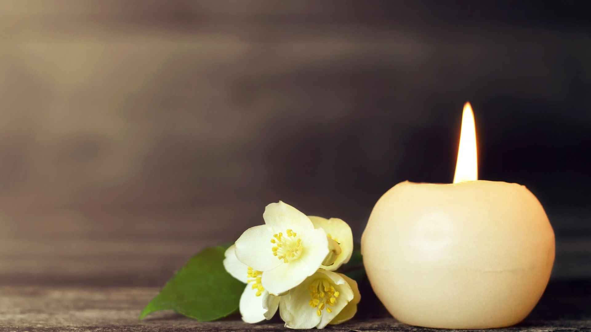 Memorial 桌布 壁紙 — 在質樸的木質表面上，一支點燃的奶油色蠟燭旁邊放著精緻的白色茉莉花。