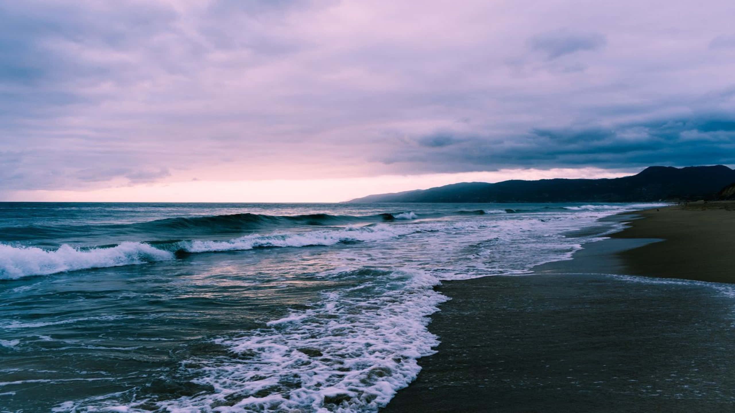 1440p Malibu 桌布 壁紙 — 在遠處山巒的襯托下，海浪拍打著深色沙灘，天空呈現憂鬱的紫色和粉紅色暮光。