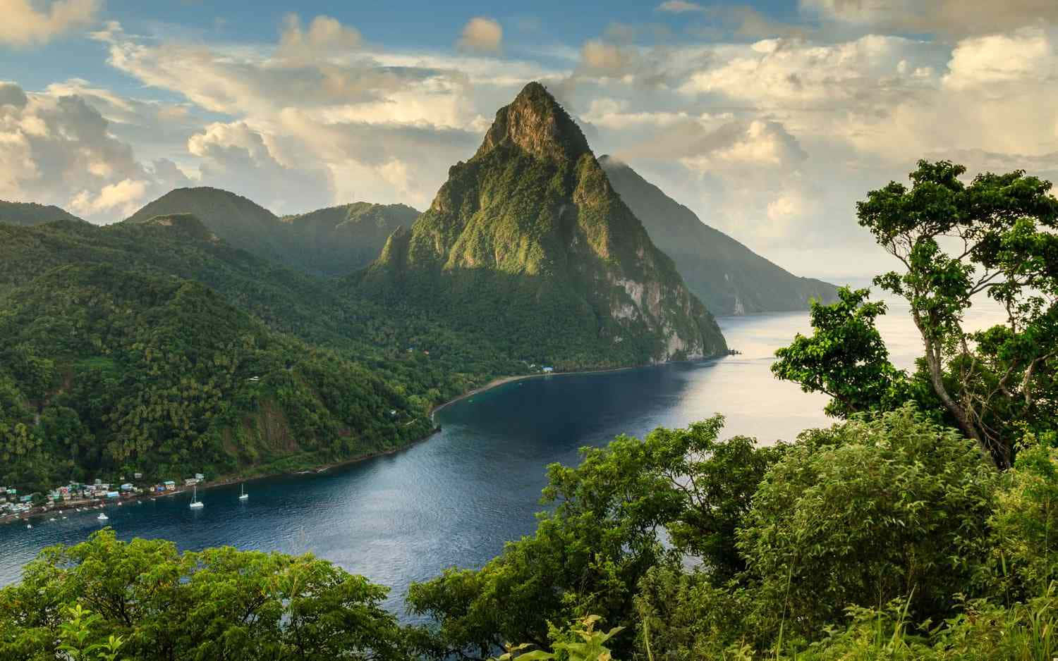 Caribbean 桌布 壁紙 — 在部分多雲的天空下，一座雄偉的綠色山峰從平靜的藍色海灣中陡峭升起，前景有茂密的樹葉。