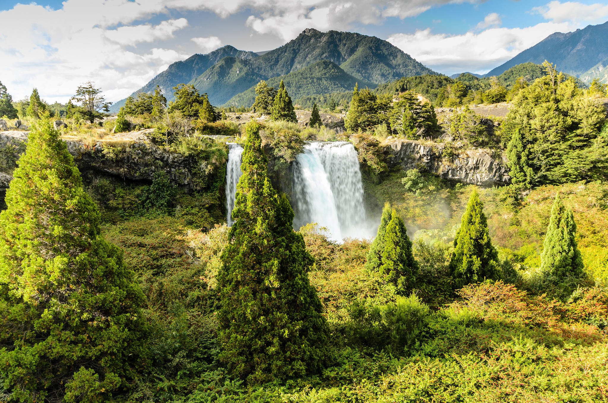 Chile 桌布 壁紙 — 在部分多雲的天空下，一道雄偉的瀑布從岩石懸崖上傾瀉而下，周圍環繞著茂密的綠樹和高聳的山脈。