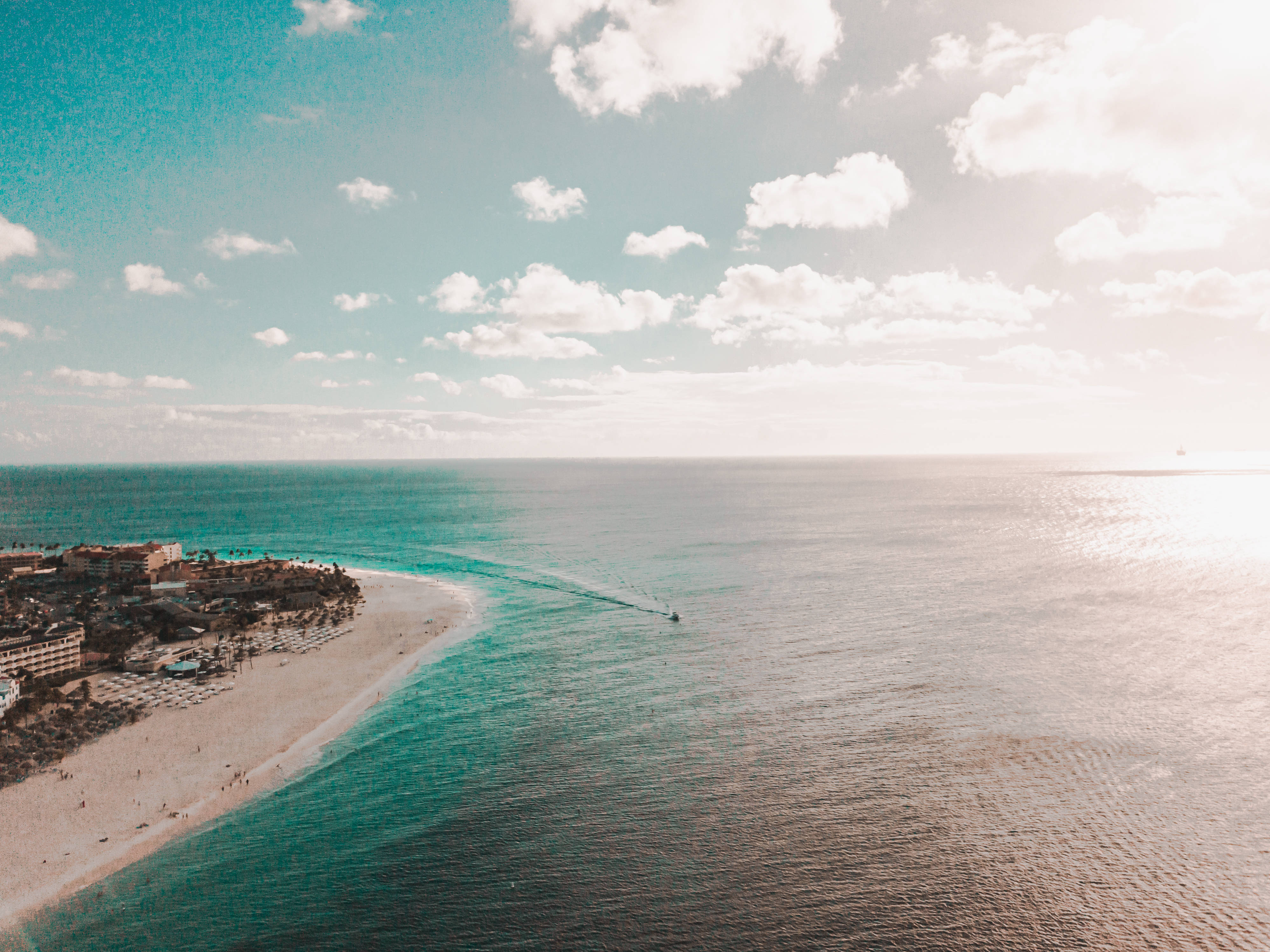 Aruba 桌布 壁紙 — 在部分多雲的天空下，彎曲的藍綠色海岸線、海灘度假村和一艘在海洋中留下白色尾跡的快艇的鳥瞰圖。