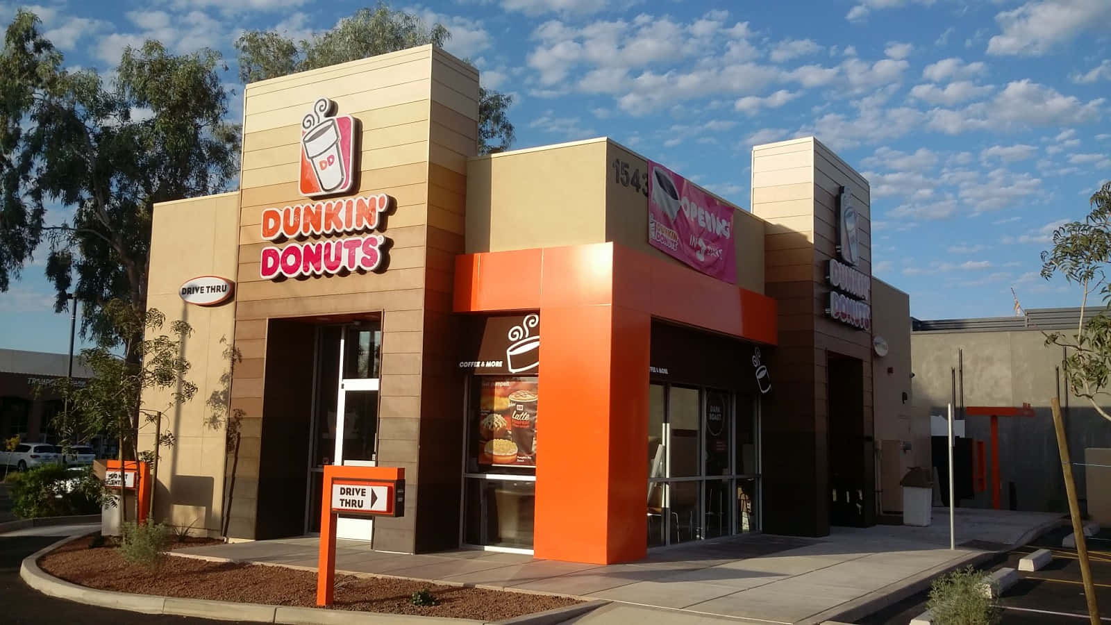 Dunkin Donuts 桌布 壁紙 — 在部分多雲的藍天下，一間亮橙色和米色的Dunkin' Donuts店舖外觀，有樹木和得來速標誌。