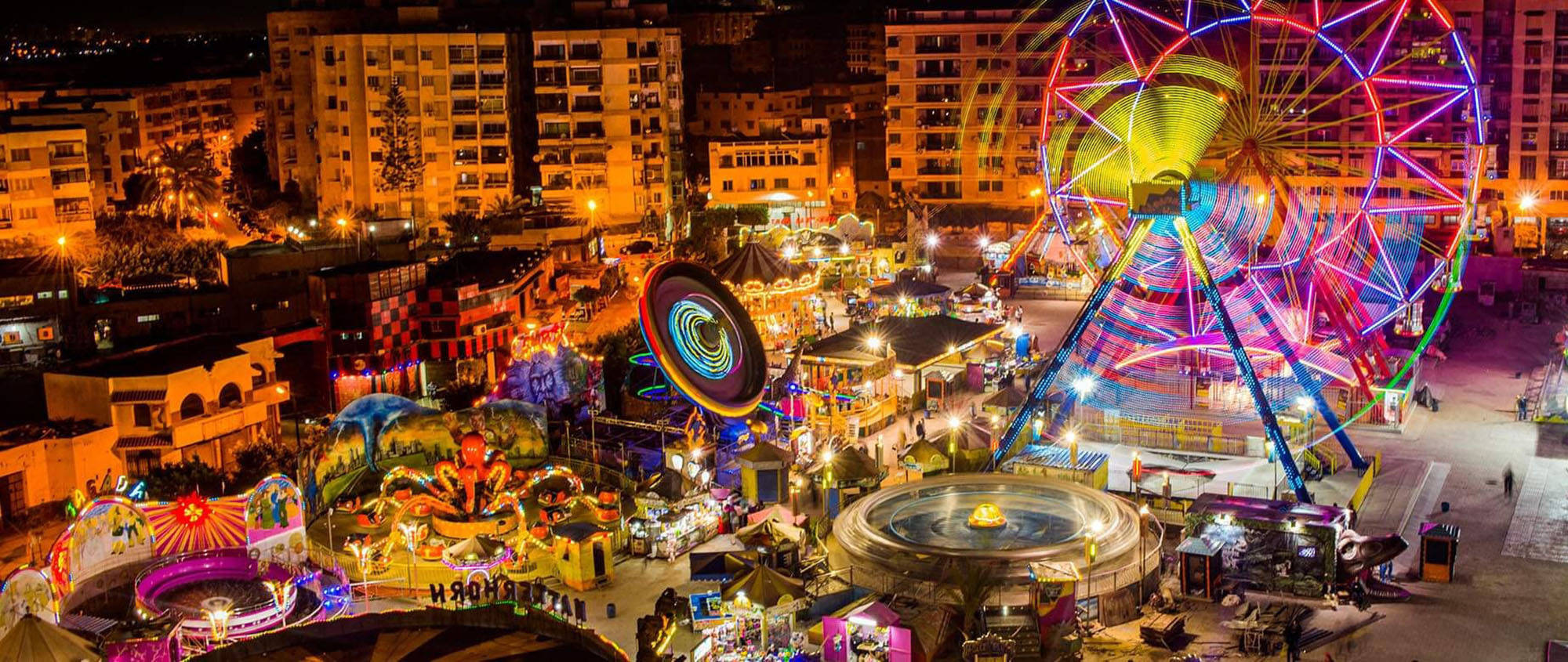 Alexandria 桌布 壁紙 — 在都市燈光下，一個令人眼花撩亂的夜間遊樂園，擁有霓虹燈照亮的遊樂設施和發光的景點。