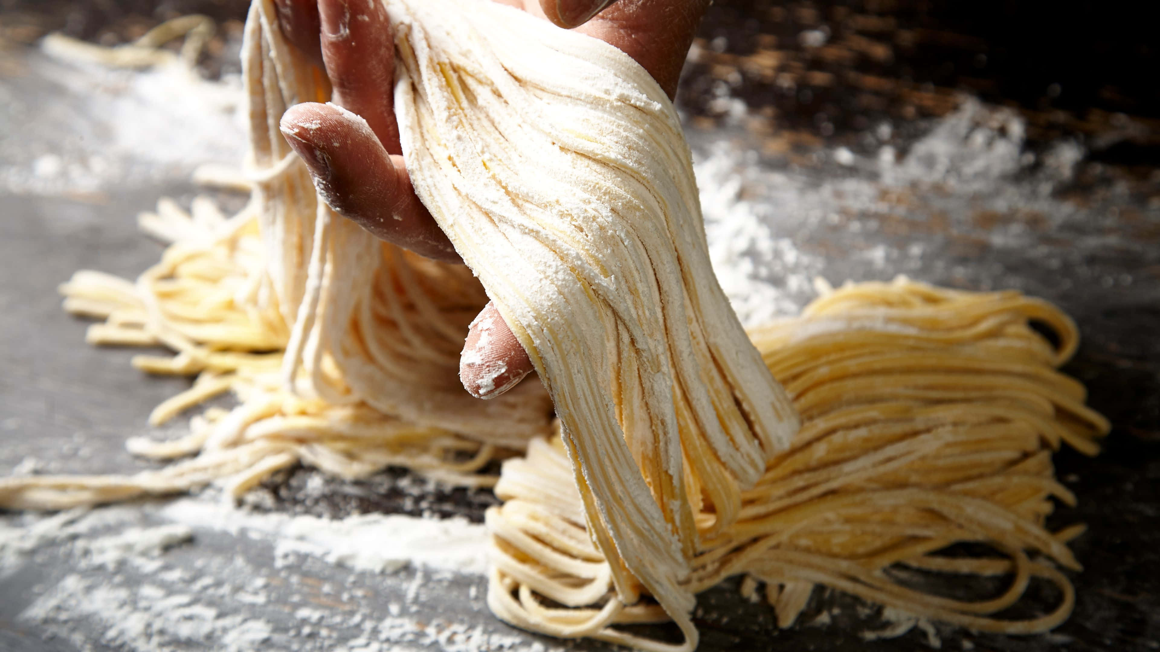Noodles 桌布 壁紙 — 在鄉村風格的廚房環境中，一隻手從撒了麵粉的木質表面上拉出新鮮的手工義大利麵條。