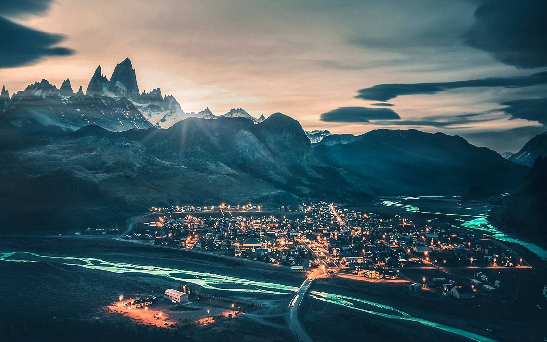 Patagonia 桌布 壁紙 — 在鋸齒狀的雪山峰頂和陰沉天空下，一座山城在黃昏時分閃爍著燈光的鳥瞰圖。
