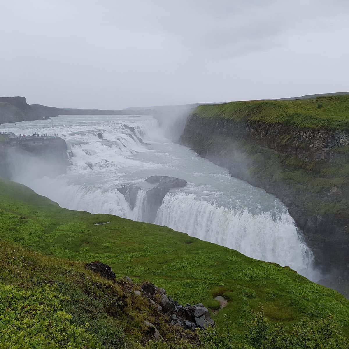 Iceland 桌布 壁紙 — 在陰天之下，一個巨大的瀑布穿過兩旁長滿翠綠懸崖的深峽谷，強力傾瀉而下。