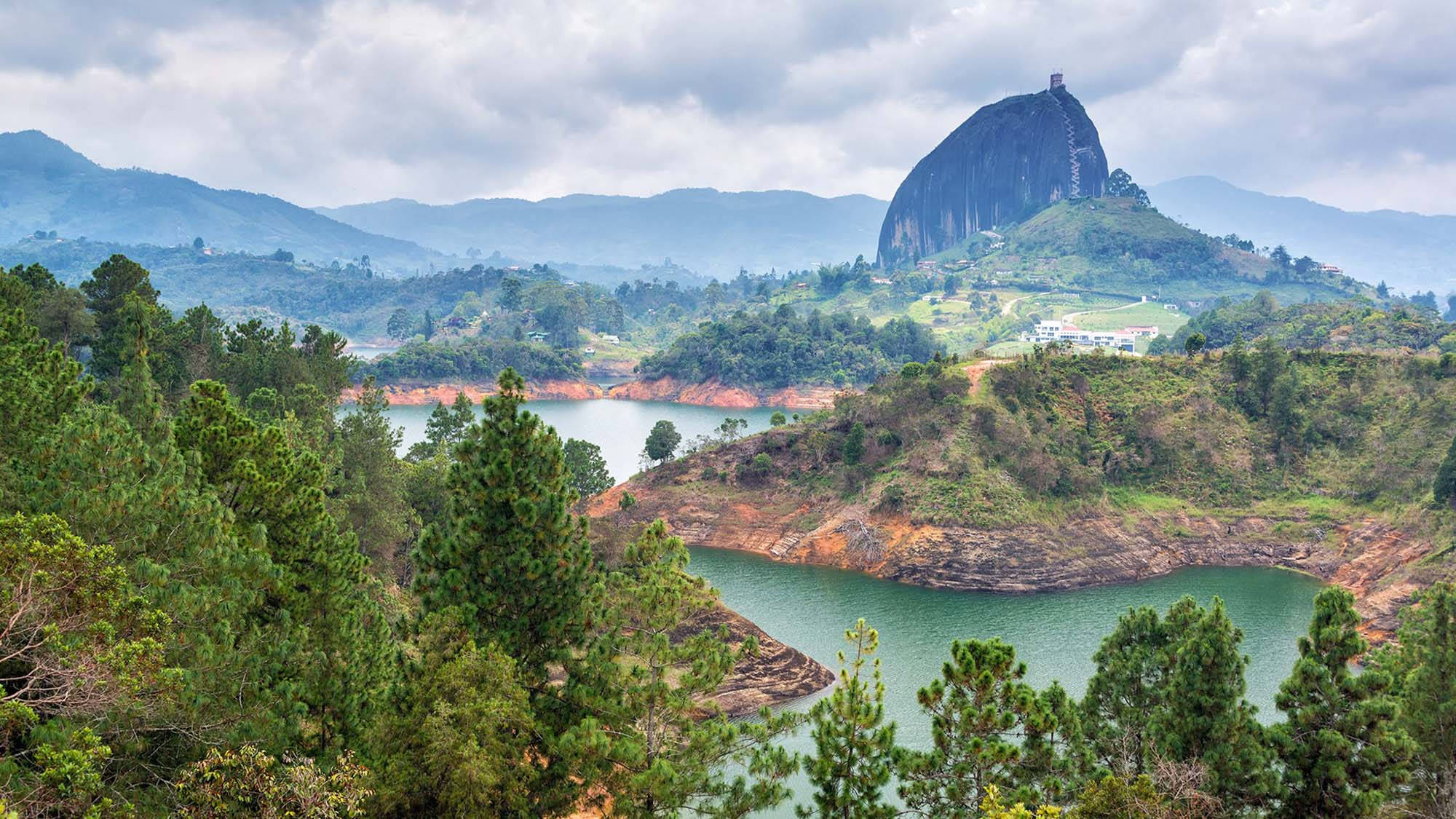 Medellin 桌布 壁紙 — 在陰天之下，一片廣闊的綠色湖泊環繞著巨大的岩石巨石，前景是森林覆蓋的山丘。