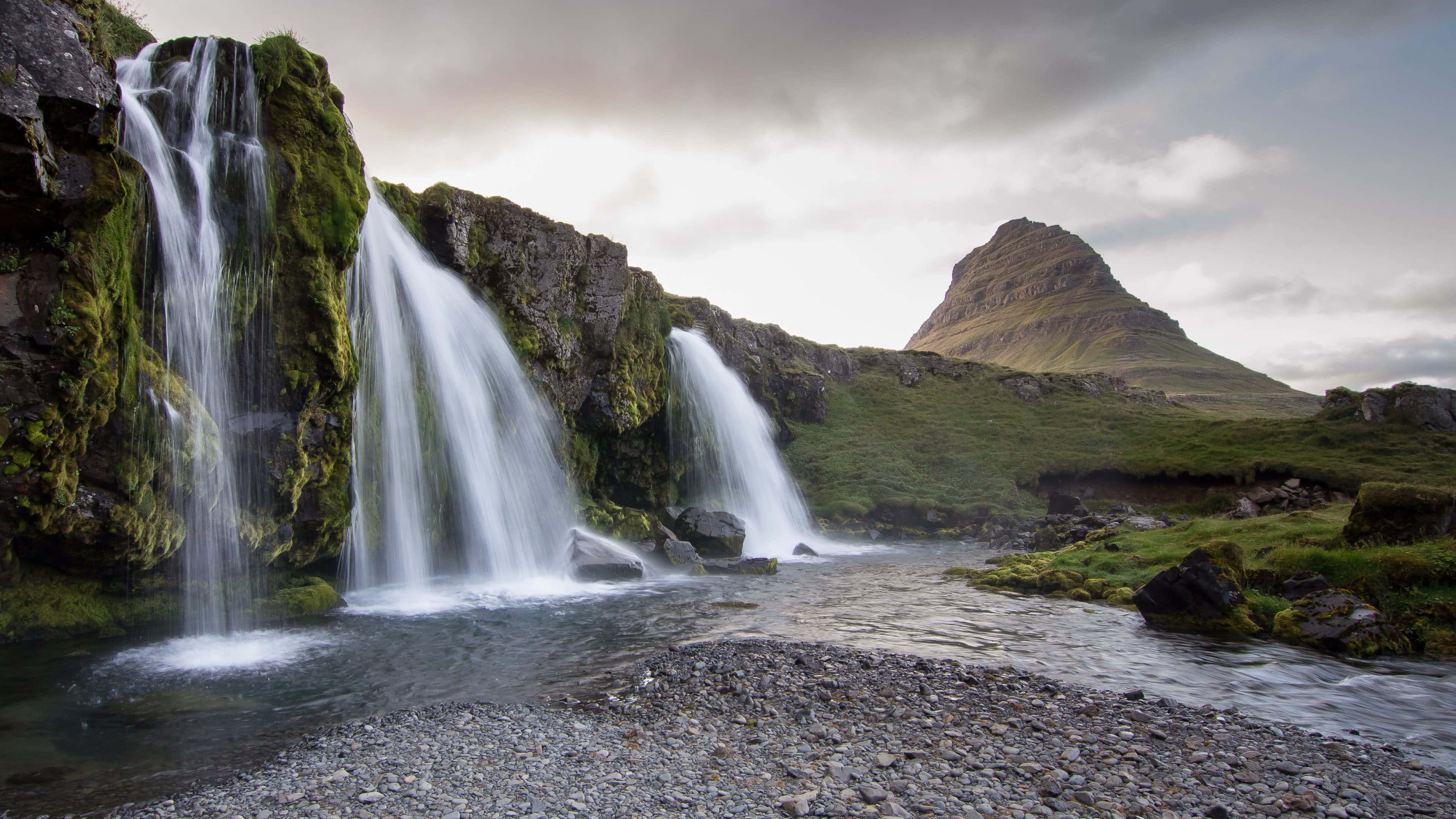 Iceland Desktop 桌布 壁紙 — 在陰天之下，一道寧靜的瀑布從長滿苔蘚的岩石傾瀉到卵石溪流中，遠處有山巒。