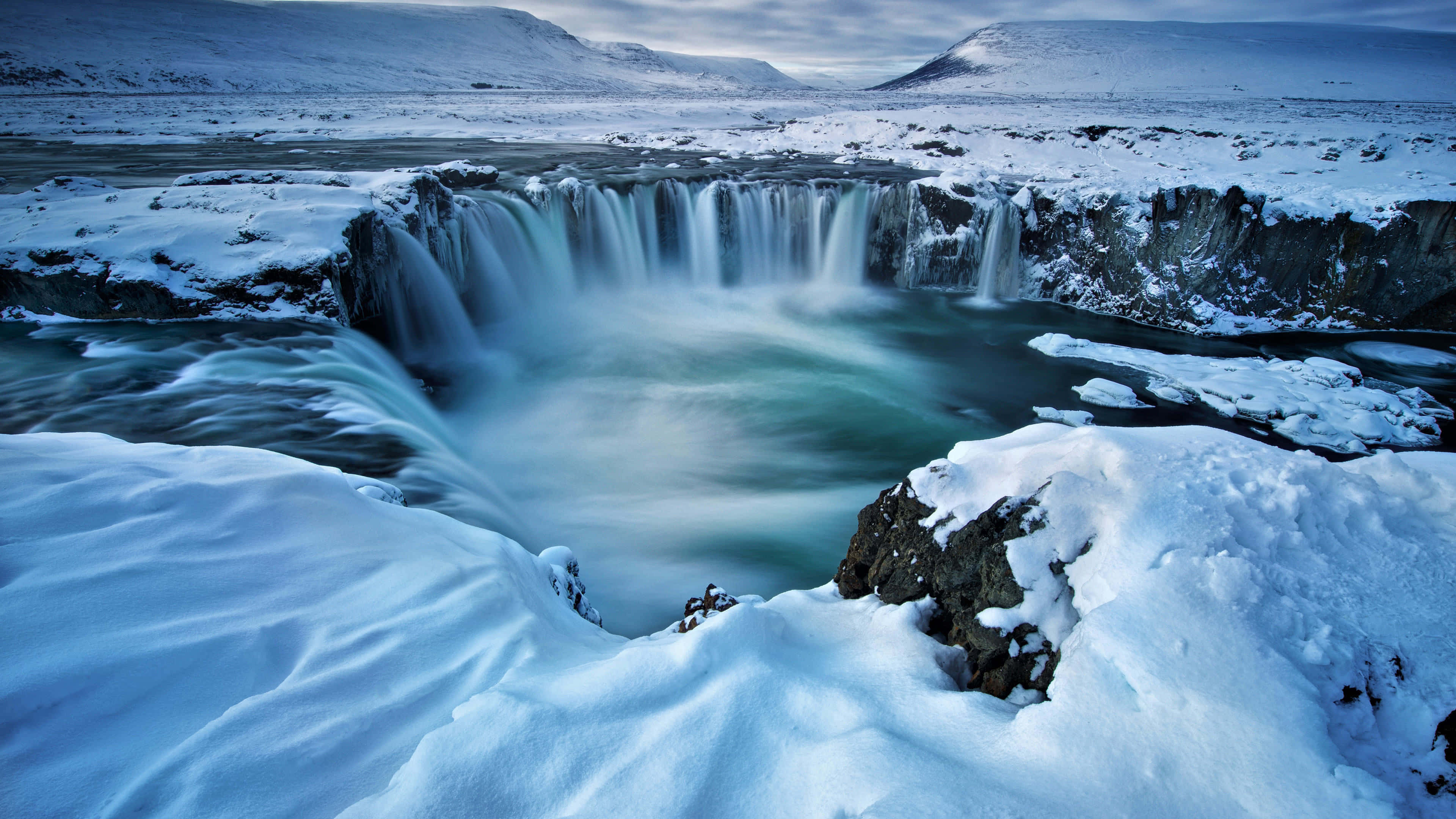 Iceland Desktop 桌布 壁紙 — 在陰天之下，一道雄偉的冬季瀑布從白雪覆蓋的懸崖傾瀉而下。