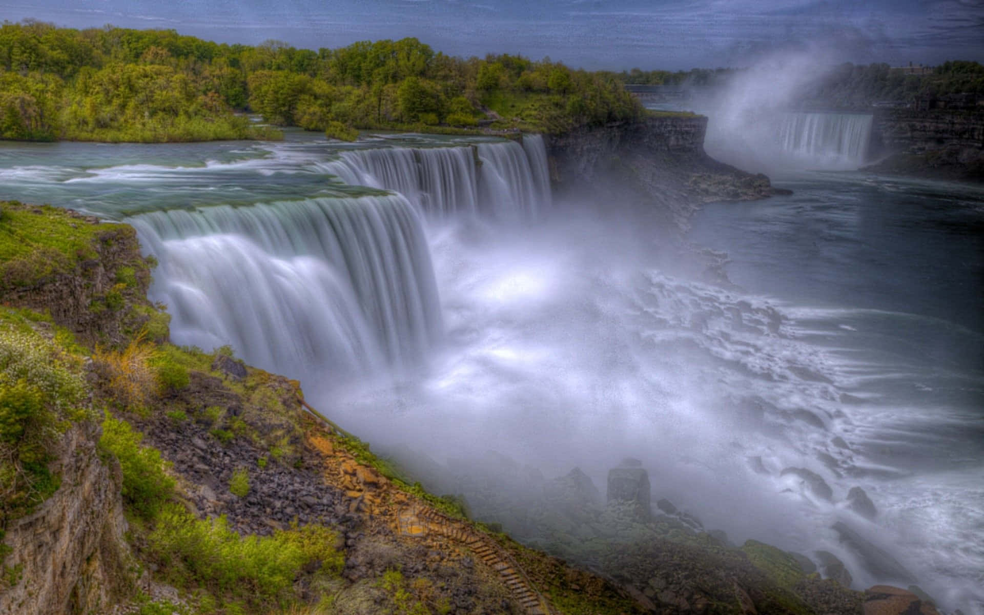 720p Niagara Falls 桌布 壁紙 — 在陰天之下，一道雄偉的寬闊瀑布從岩石懸崖傾瀉而下，流入被茂密綠色森林環繞的霧氣瀰漫的河流。