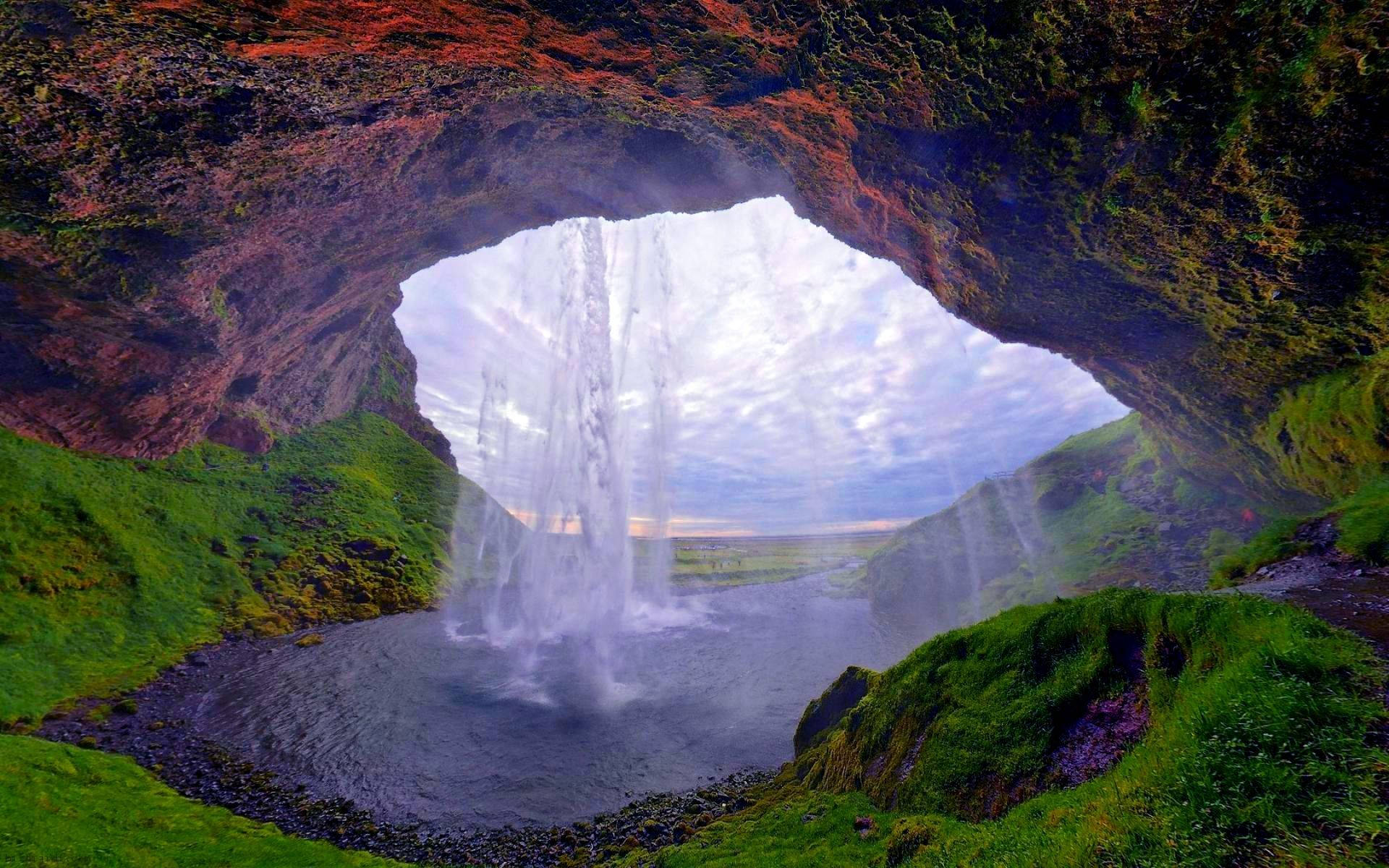 瀑布流經長滿苔蘚的岩石拱門，周圍是鬱鬱蔥蔥的綠色景觀。 — 瀑布 GIF 壁紙
