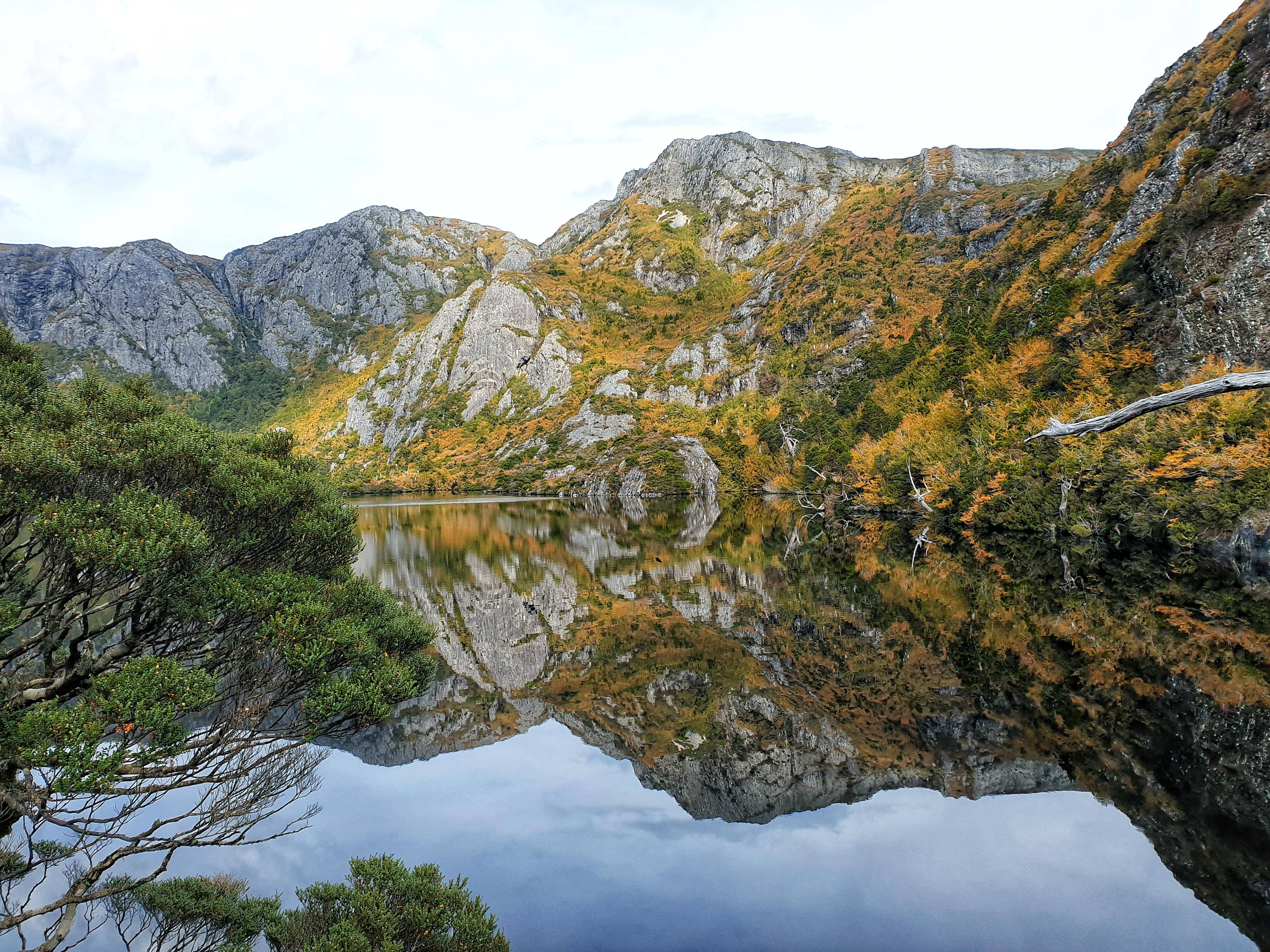自然 桌布 壁紙 — 在陰天之下，寧靜的山中湖泊映照著崎嶇的山峰和秋葉。