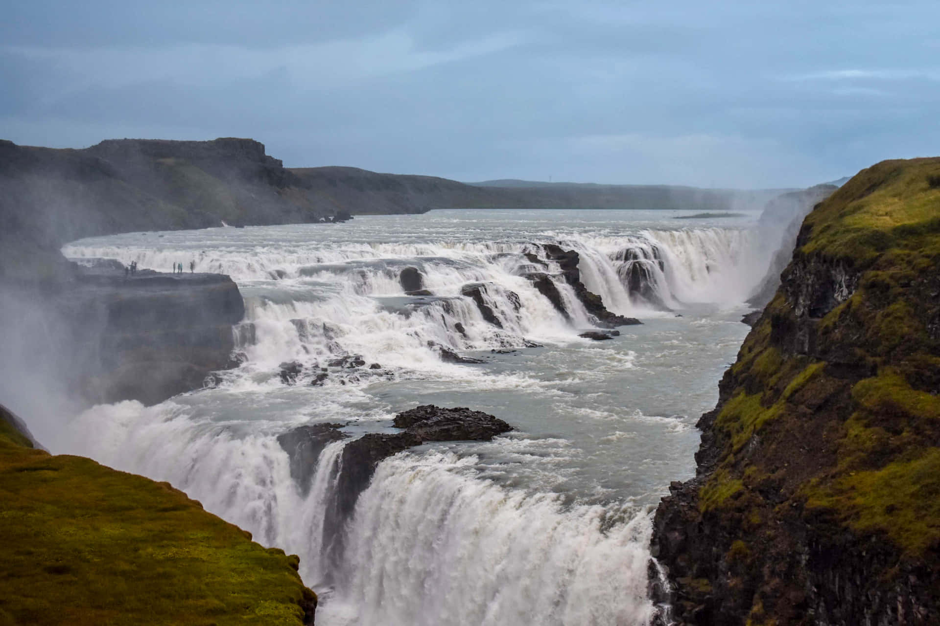 Iceland 桌布 壁紙 — 在陰天之下，巨大的瀑布從長滿苔蘚的懸崖傾瀉而下，翻騰的水面升起薄霧。