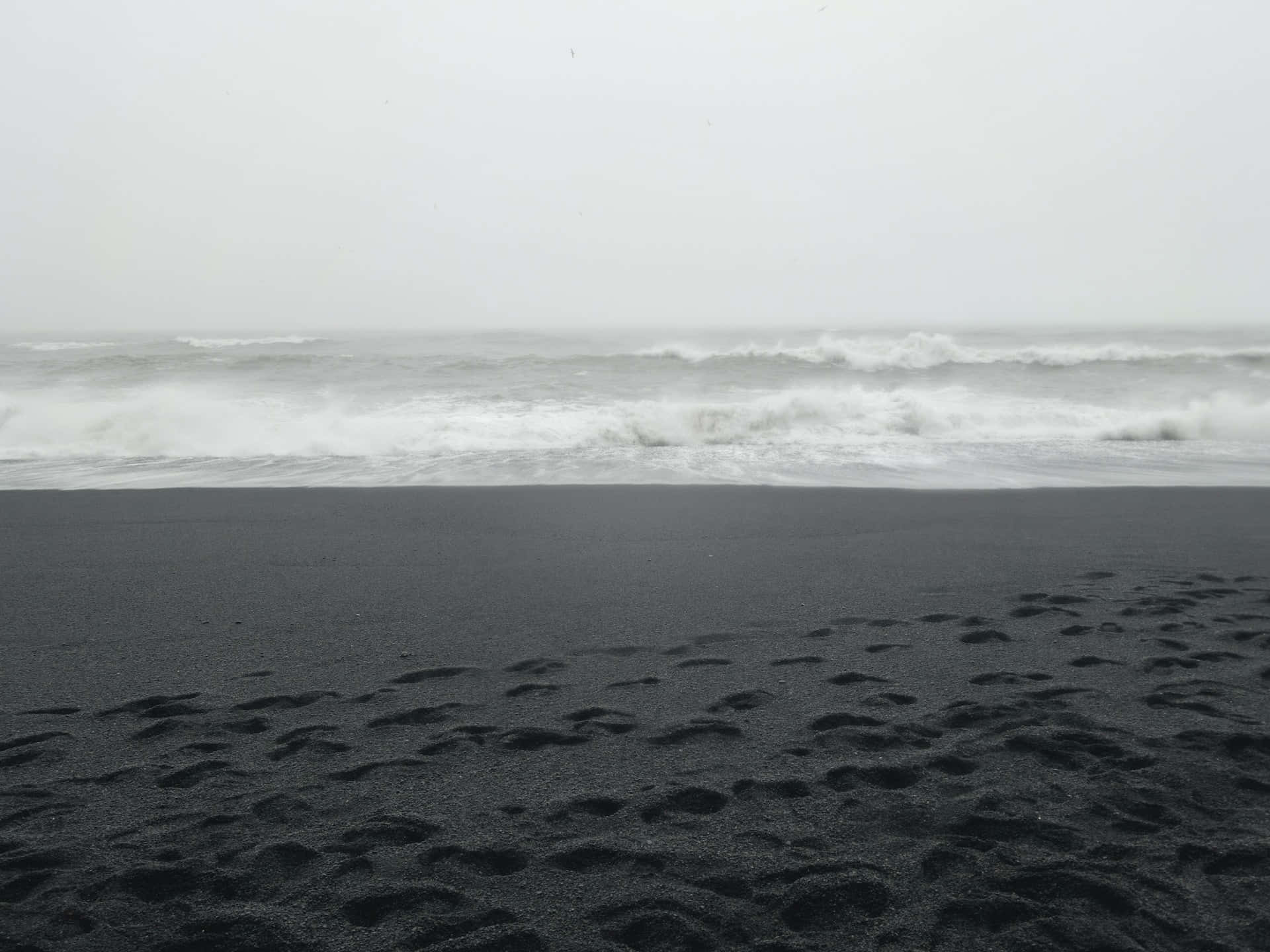 Black And White Beach 桌布 壁紙 — 在陰天之下，海浪拍打著黑色的火山沙灘，前景有腳印。