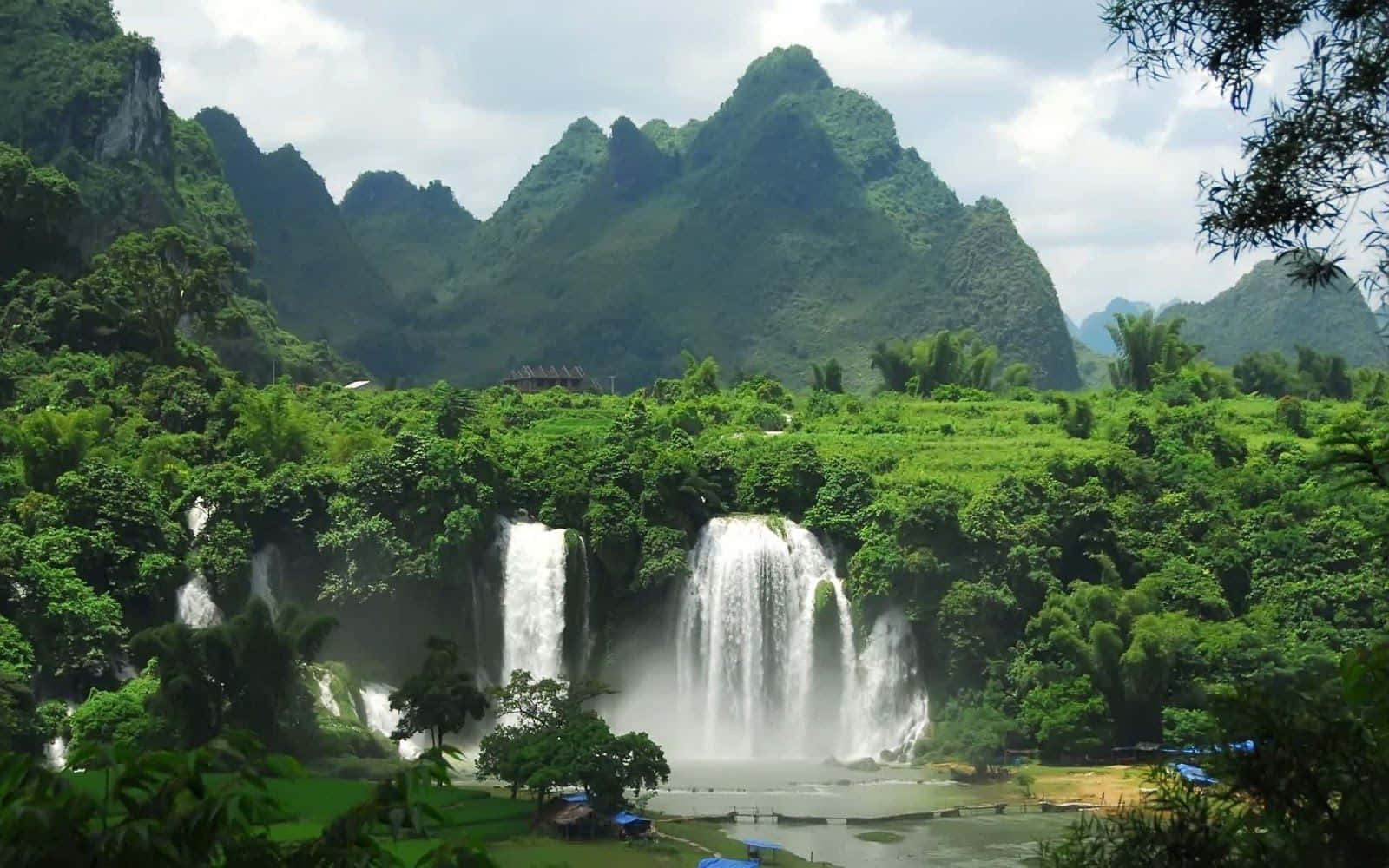 風景 桌布 壁紙 — 在陰天之下，雄偉的瀑布穿過茂密的綠色叢林，背景是高聳的喀斯特山脈。