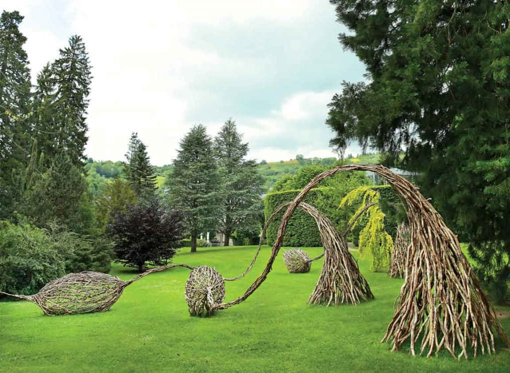 Land Art 桌布 壁紙 — 在陰天之下，高大的常綠樹環繞著一片充滿活力的綠色草坪，奇特的柳樹雕塑在其上拱起和彎曲。