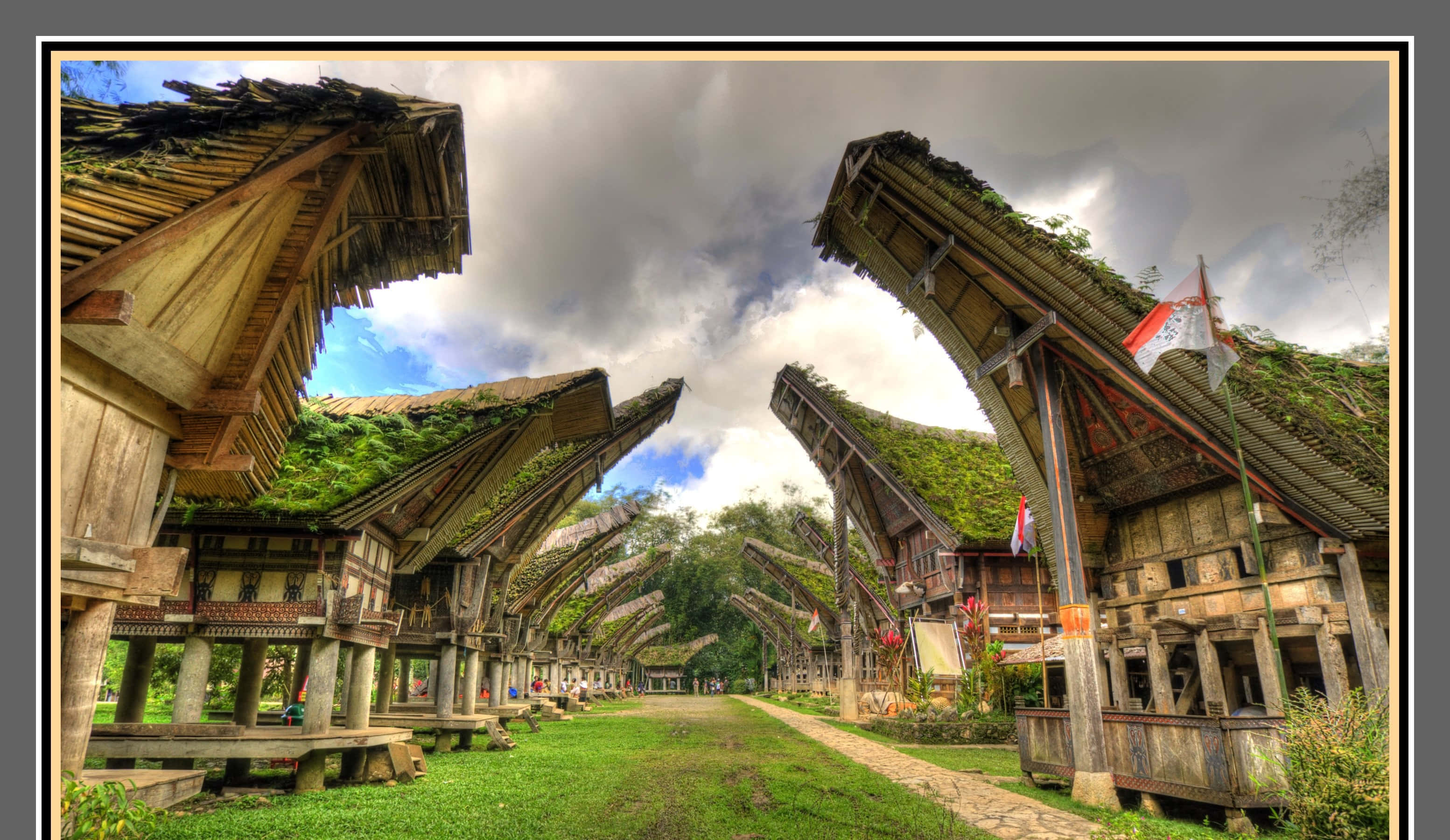 Architecture 桌布 壁紙 — 在陰天天空下，傳統高腳屋以陡峭茅草屋頂排列在草地小徑旁的戲劇性低角度視圖。