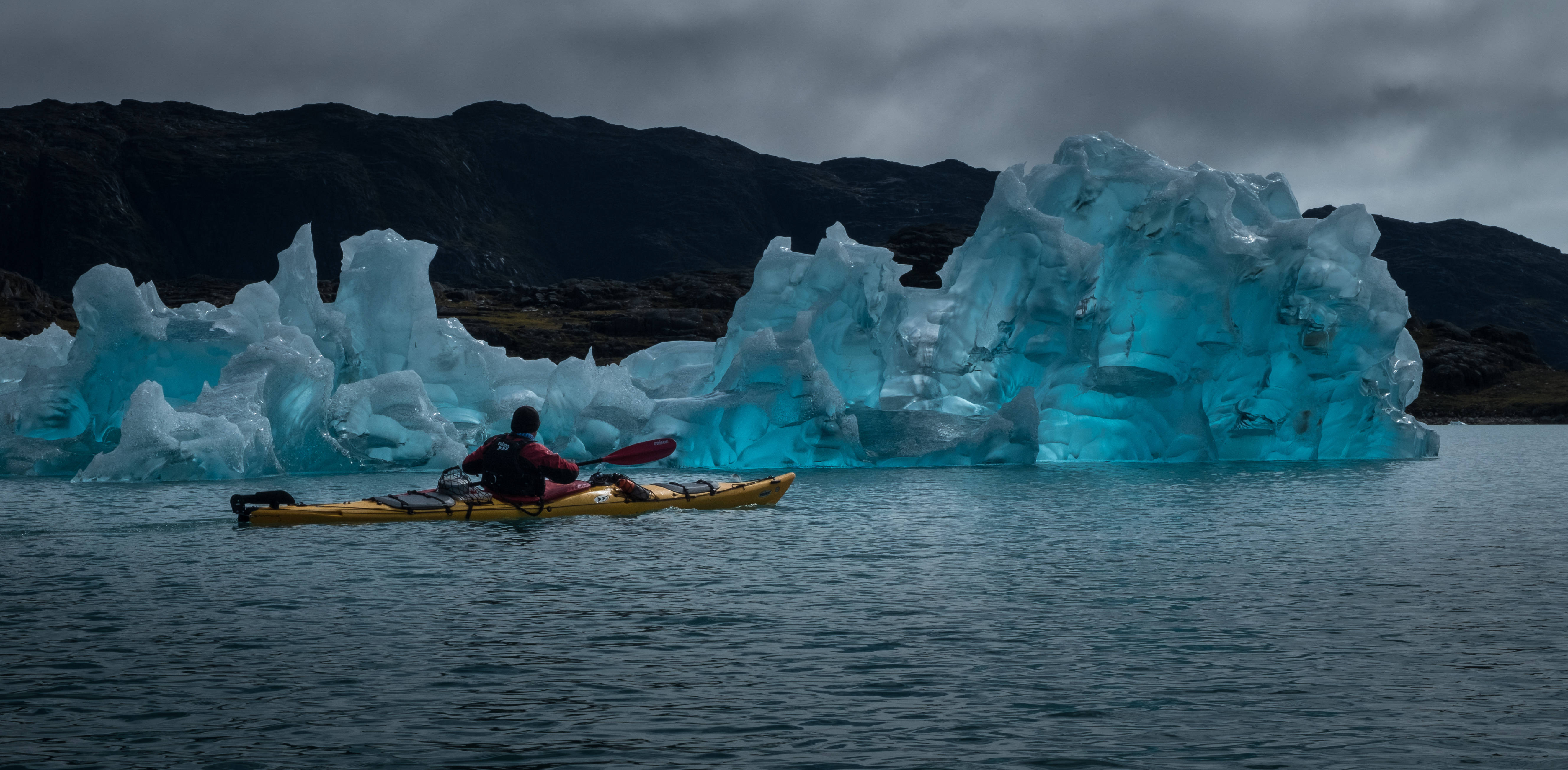Greenland 桌布 壁紙 — 在陰暗多雲的天空下，一名皮划艇運動員在巨大的發光藍色冰山附近划著黃色皮划艇。