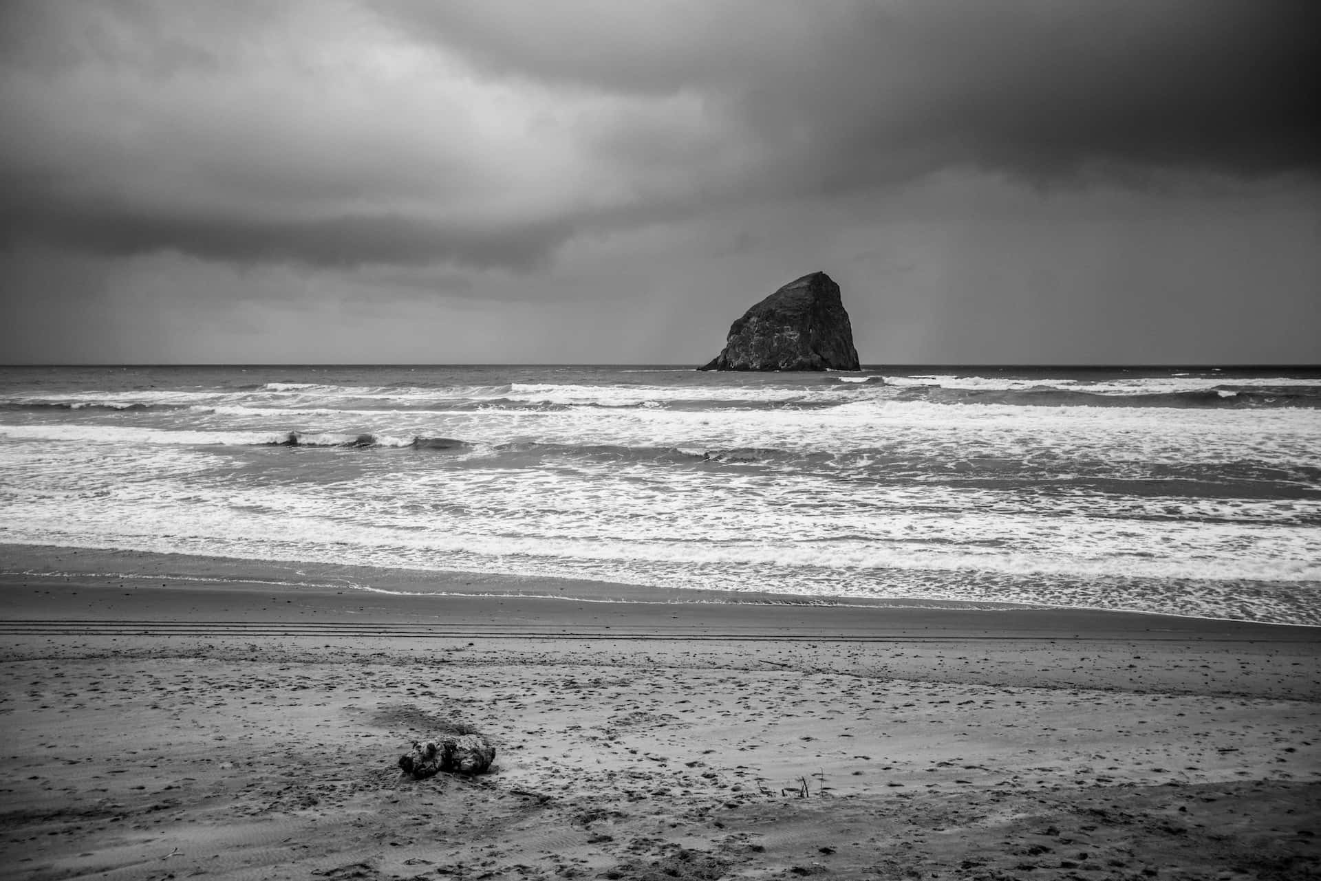 Black And White Beach 桌布 壁紙 — 在陰沉密佈的天空下，一座孤獨的海蝕柱從湍急的海水中升起的單色海岸場景。
