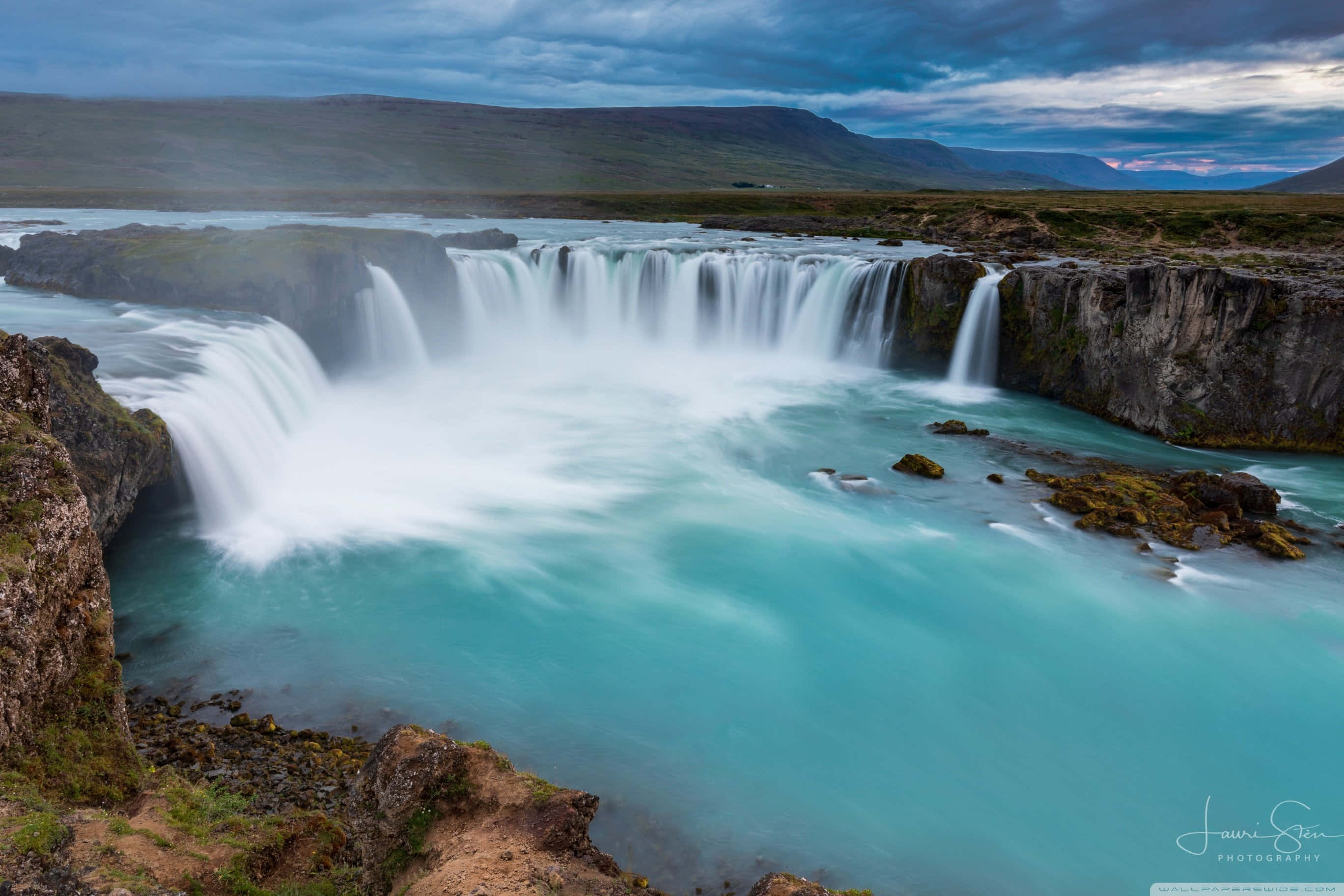 Iceland Desktop 桌布 壁紙 — 在陰沉的暮色天空下，一道雄偉的寬闊瀑布傾瀉入一個藍綠色的水池，遠方有山巒。