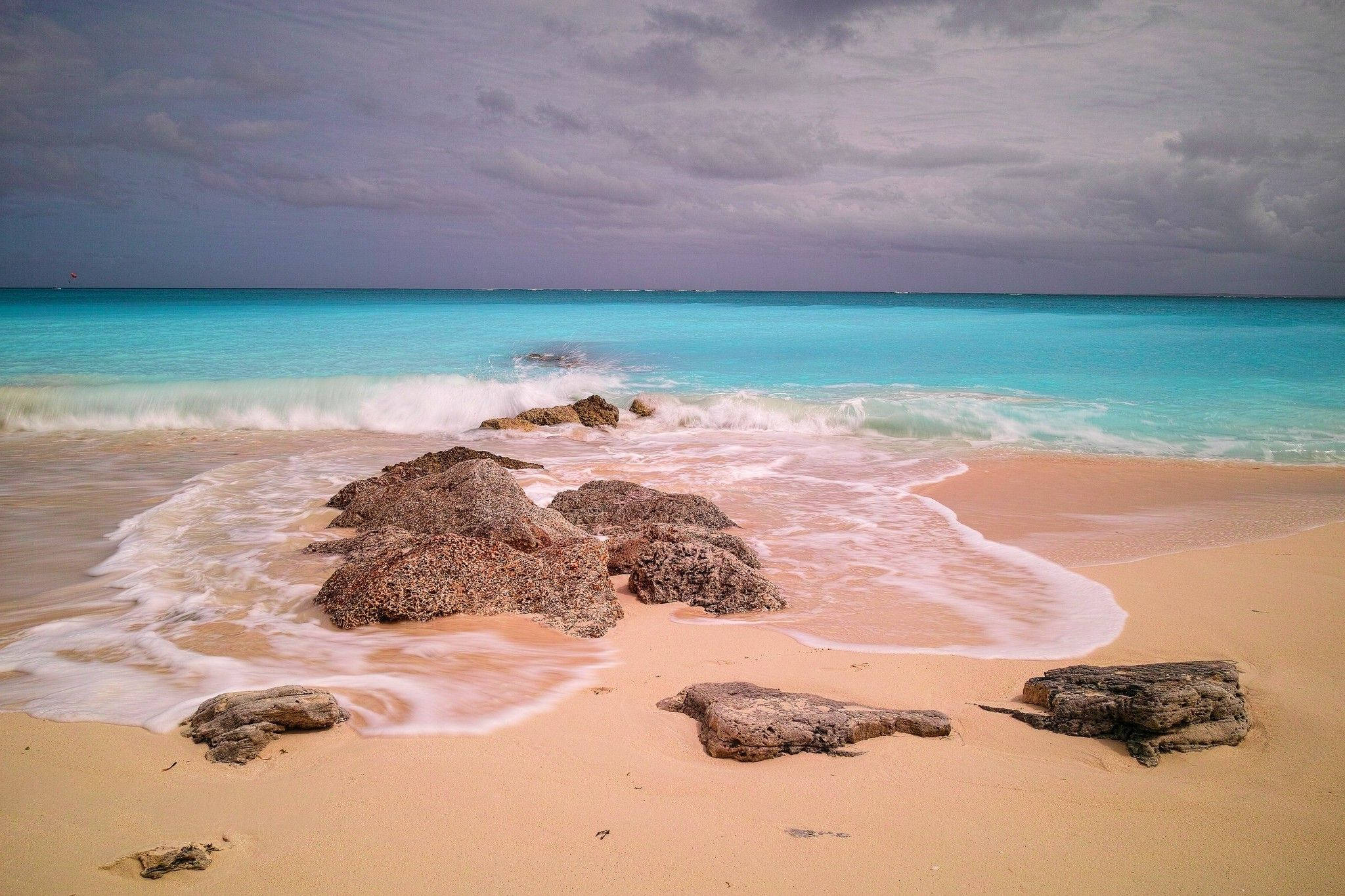 Turks And Caicos 桌布 壁紙 — 在陰沉的灰色天空下，綠松石色的海浪拍打著散佈著崎嶇岩石的沙灘。