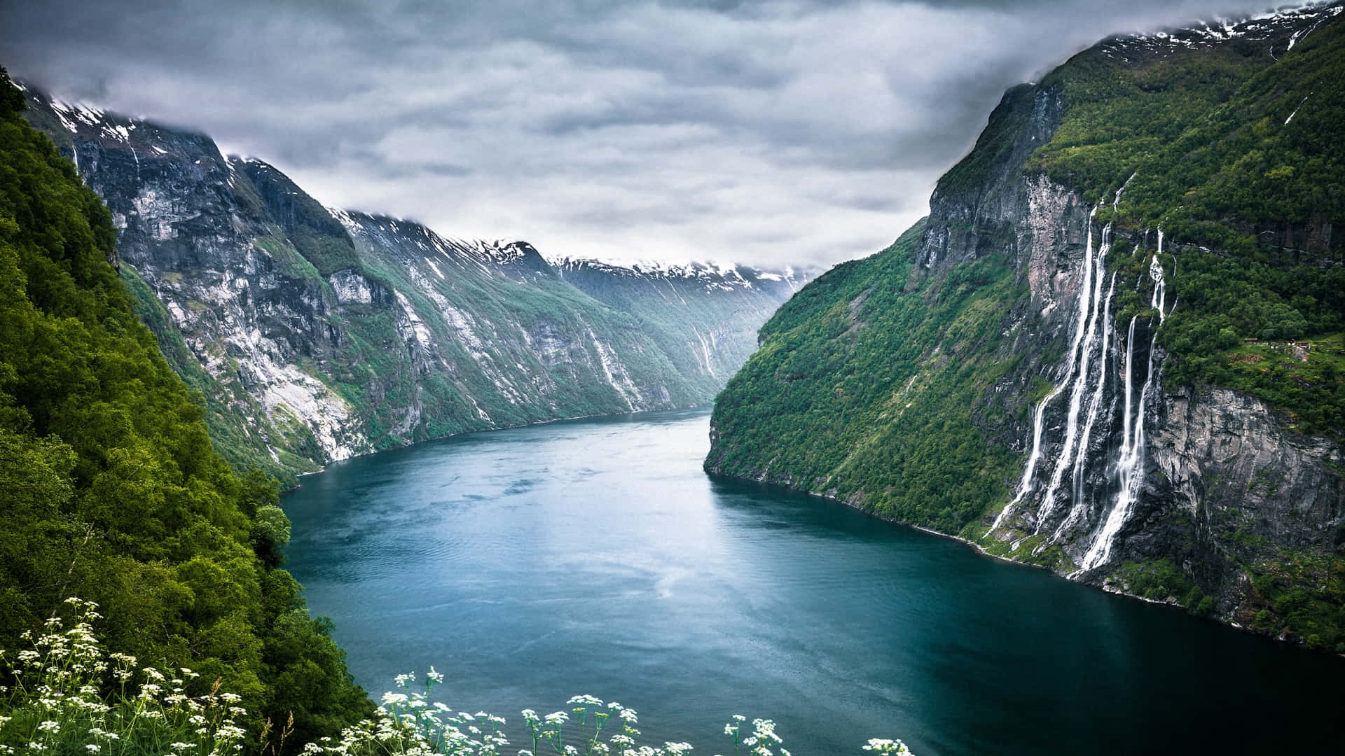 Norway 桌布 壁紙 — 在陰雲密布的天空下，一條雄偉的峽灣蜿蜒於高聳的綠色懸崖之間，懸崖上有積雪的山峰和傾瀉而下的瀑布。