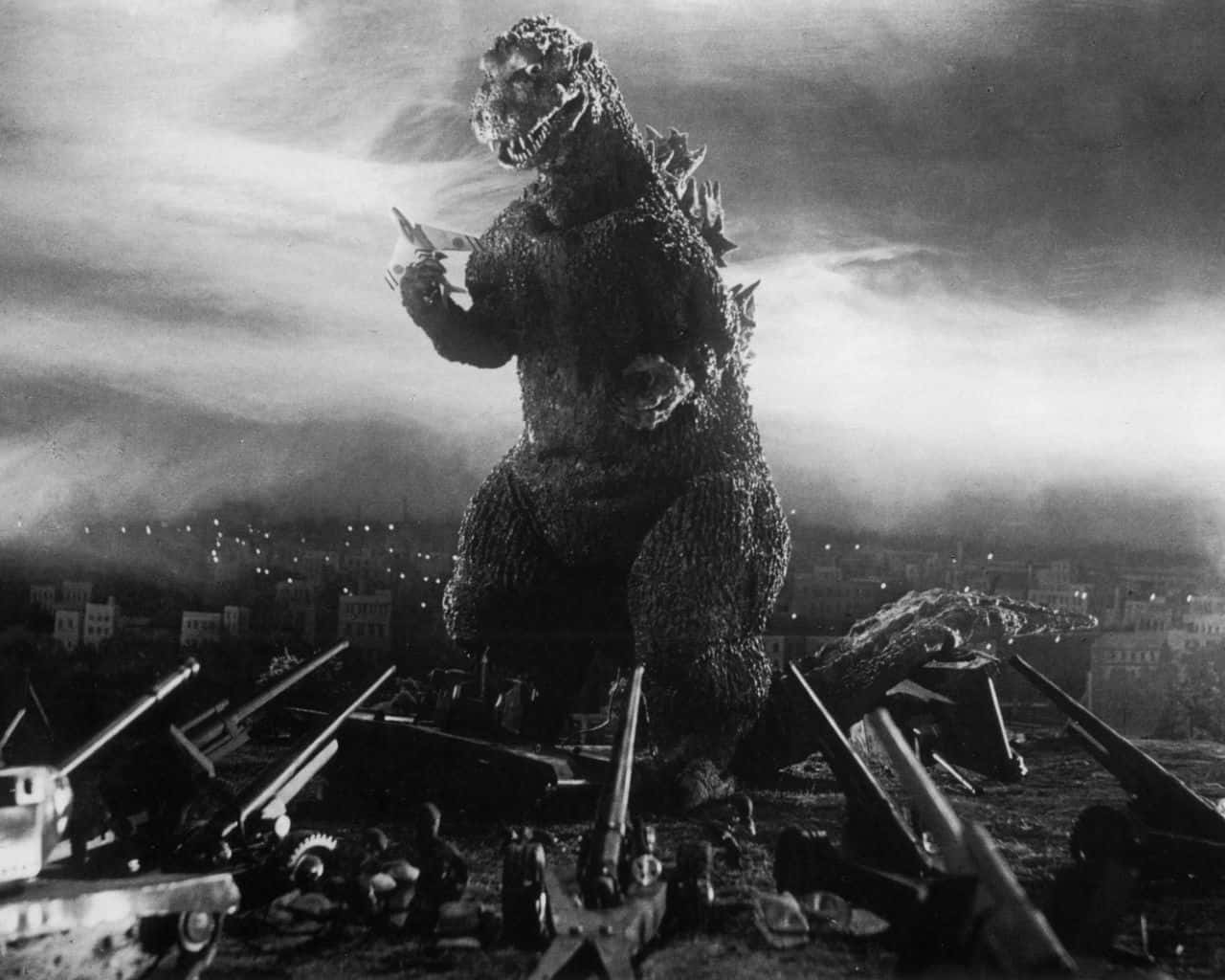 Godzilla 1954 桌布 壁紙 — 在陰雲密布的天空下，一隻高聳的哥吉拉站立於黑白城市景觀中被摧毀的坦克和瓦礫之中。