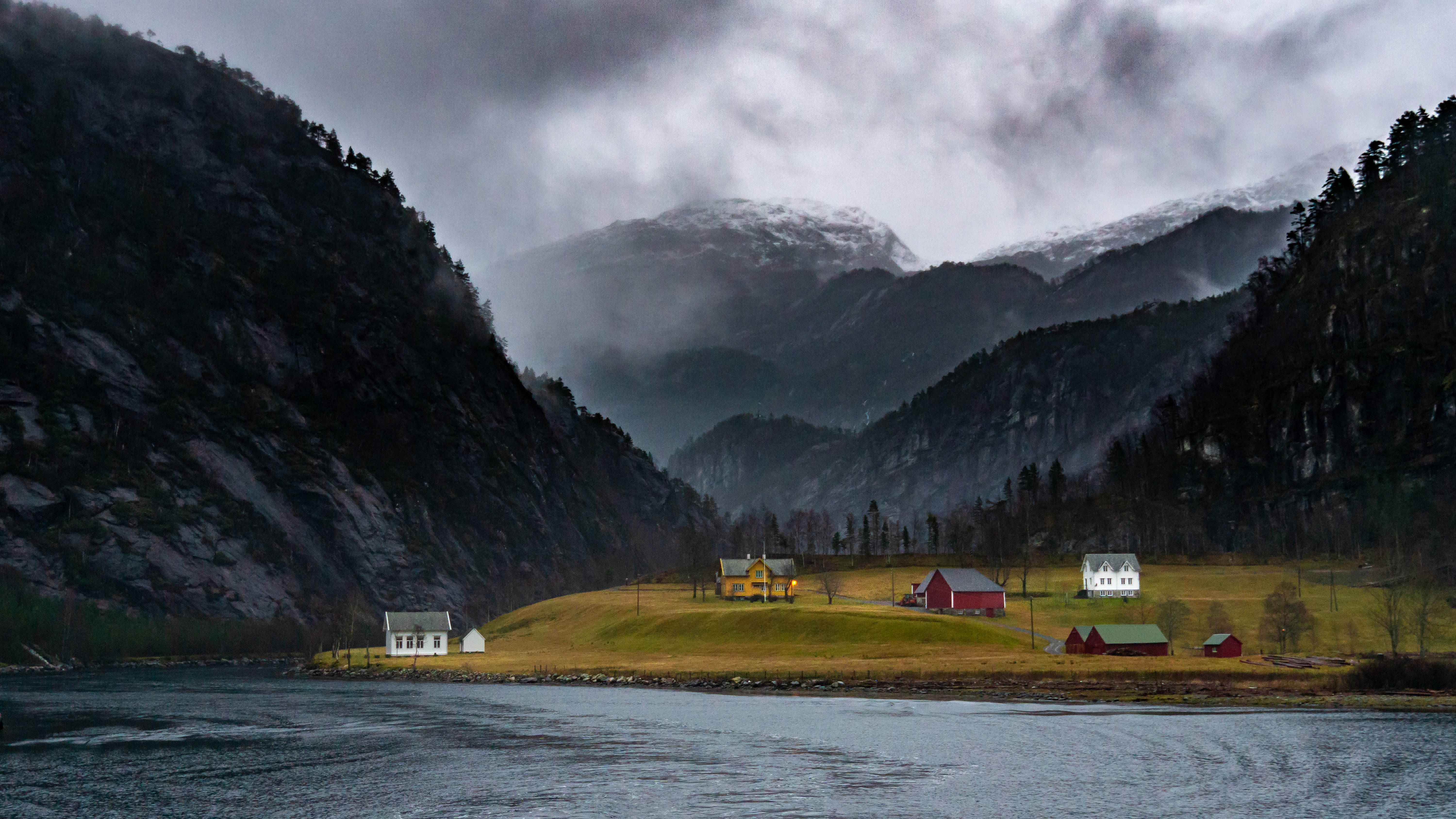 Norway 桌布 壁紙 — 在陰雲密布的天空下，霧氣繚繞的峽灣山谷中，色彩繽紛的小屋坐落在高聳的岩石山之間。