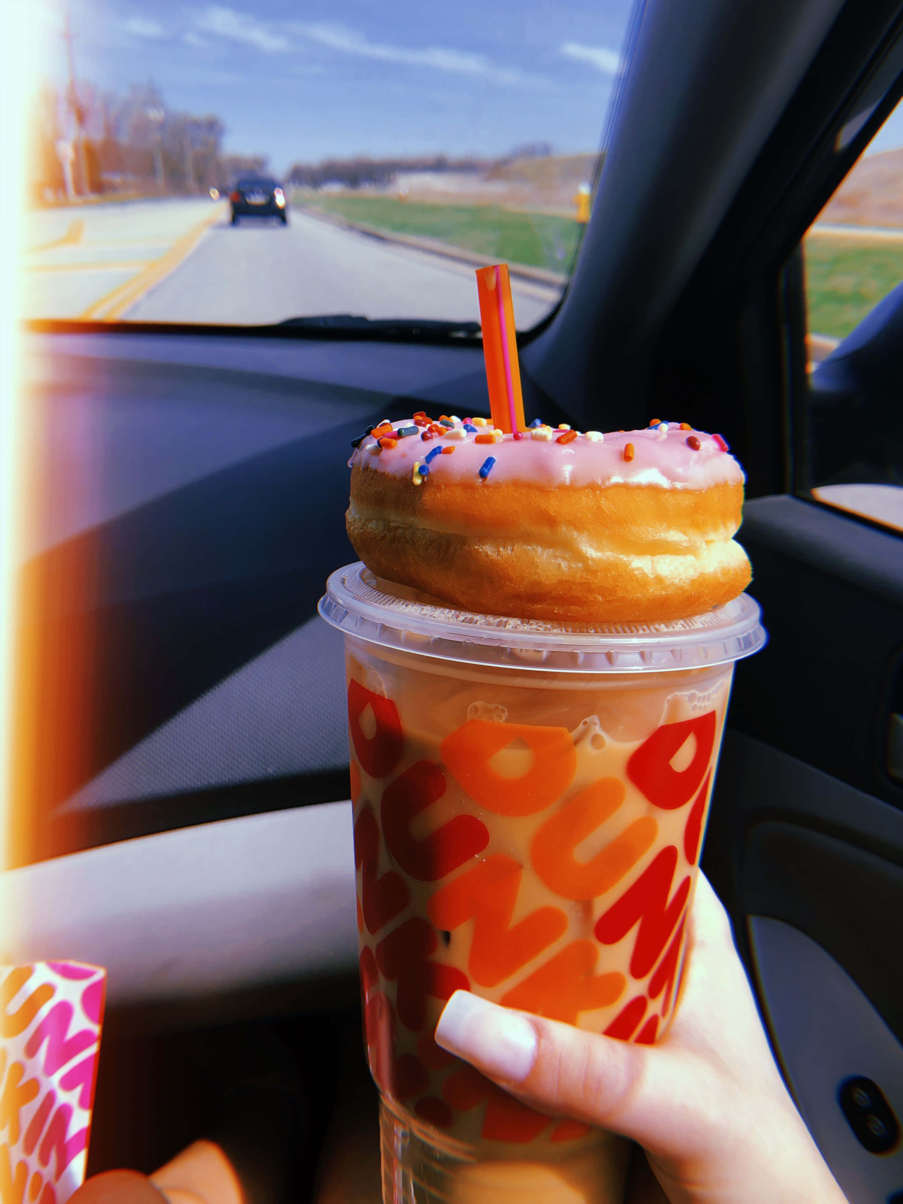 Dunkin Donuts 桌布 壁紙 — 在陽光明媚的公路旅行日，一隻手在車內拿著一杯頂部有粉紅色糖霜甜甜圈和糖粒的Dunkin'冰咖啡。