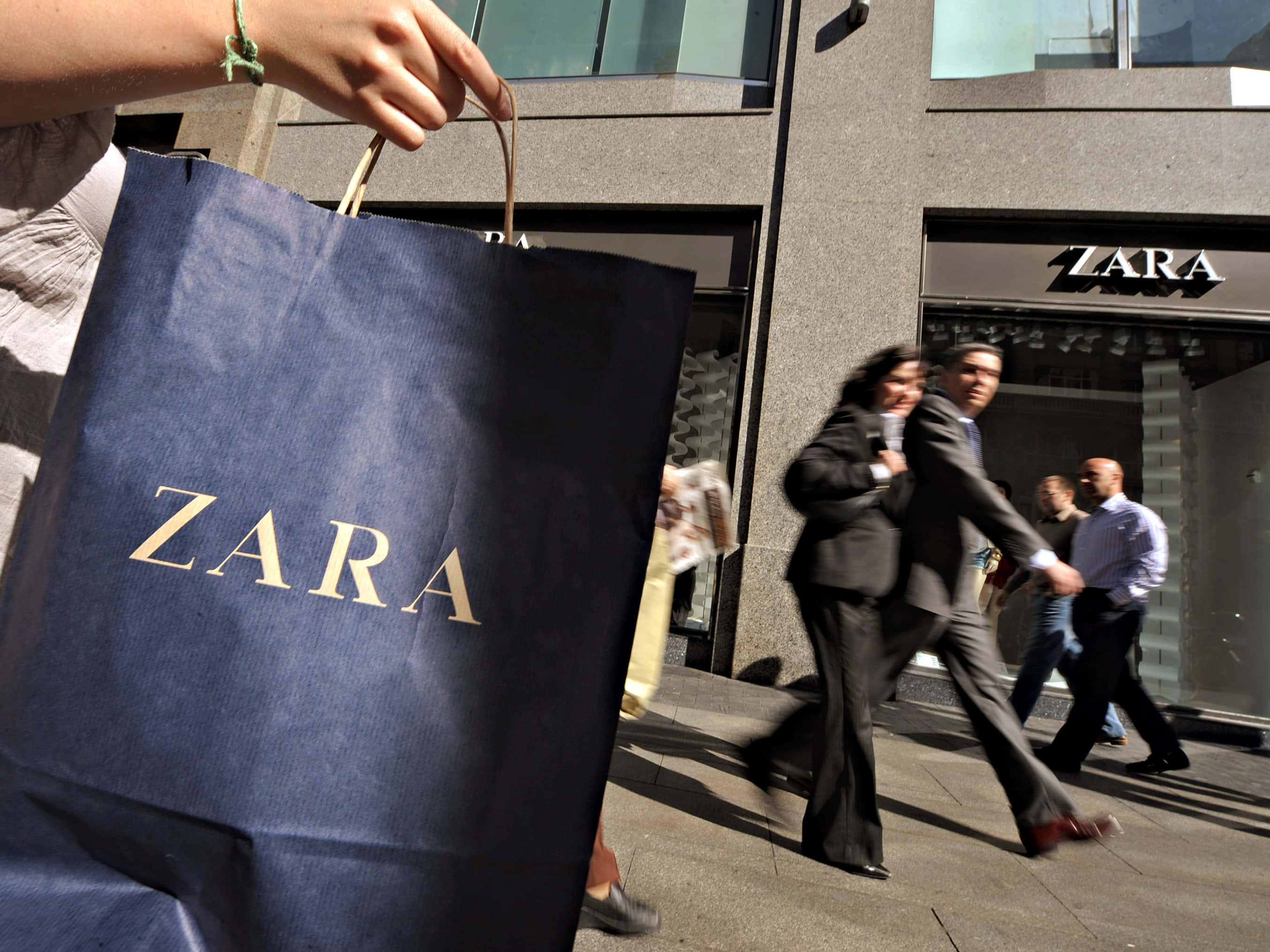 Zara 桌布 壁紙 — 在陽光明媚的城市人行道上，一名行人手持深藍色Zara購物袋，行人匆匆走過Zara商店。