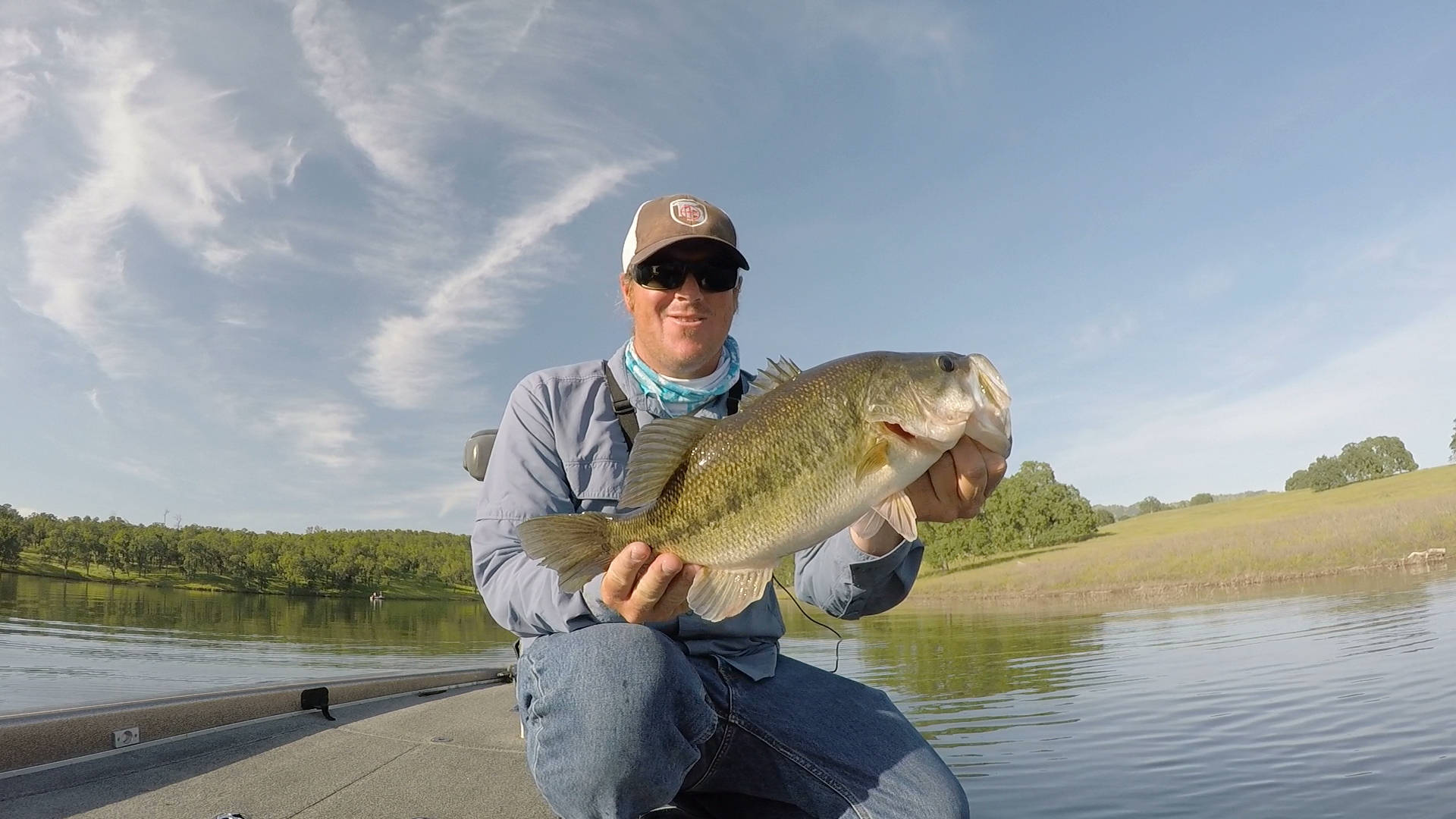 Largemouth Bass 桌布 壁紙 — 在陽光明媚的日子裡，一位微笑的釣魚人從平靜湖面上的船上自豪地舉起一條大鱸魚，背景有綠樹和藍天。